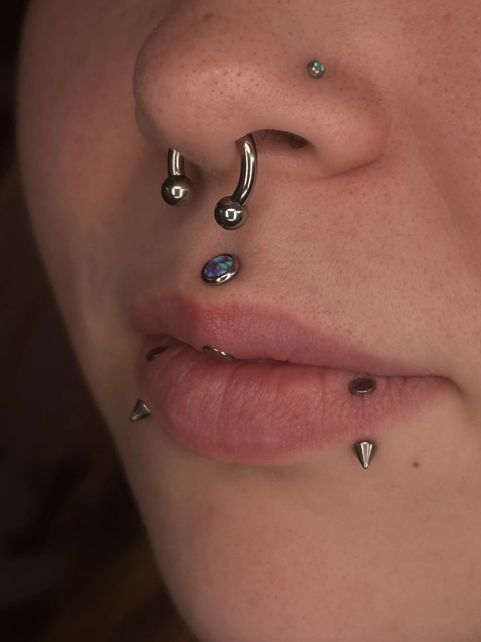 Close-up of a person's face showcasing multiple facial piercings, including a septum piercing with a curved barbell, a nostril piercing, and a labret piercing with a decorative gem. The person's skin has a natural tone with visible pores.