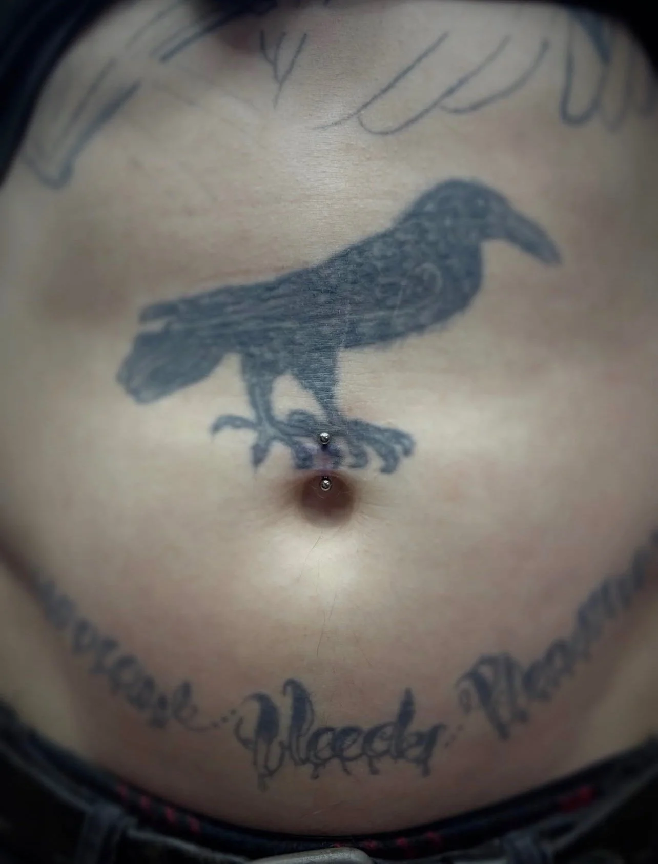 Close-up of a tattoo of a black bird, possibly a raven, on someone's chest with some text above and below it, and a piercing below the tattoo.