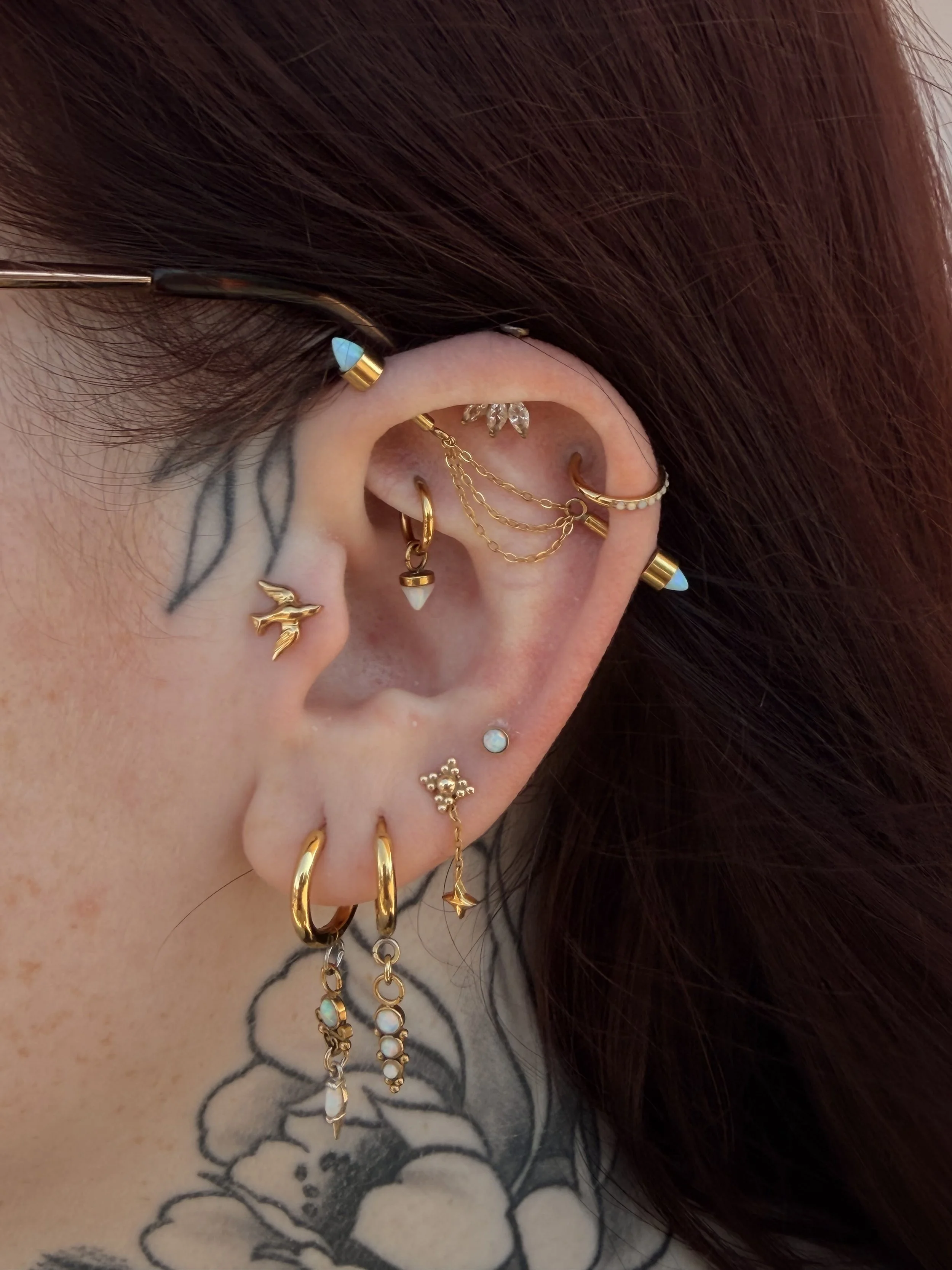 Close-up of a person's ear and neck showing numerous earrings including gold hoops, chains, and decorative studs with opal and crystal accents, and a tattoo of flowers and leaves on the neck.