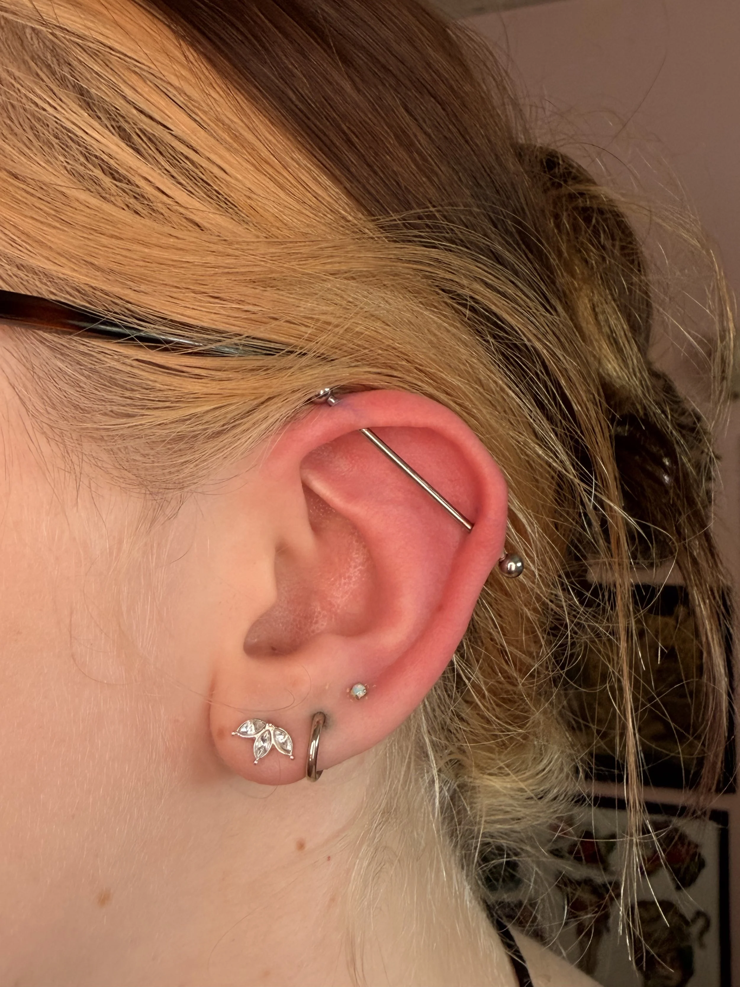 Close-up of a person's ear with multiple piercings, including a helix, lobe, and tragus, adorned with various silver earrings and jewelry, including a leaf-shaped earring and a small stud, with part of their blonde hair visible.