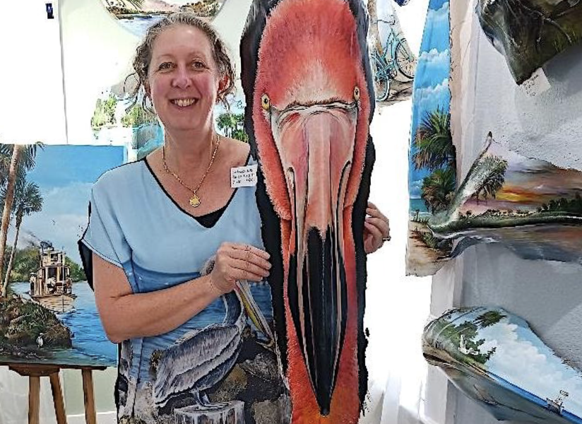 A smiling woman holding a large artwork of a flamingo near paintings of tropical scenes and a boat on water.