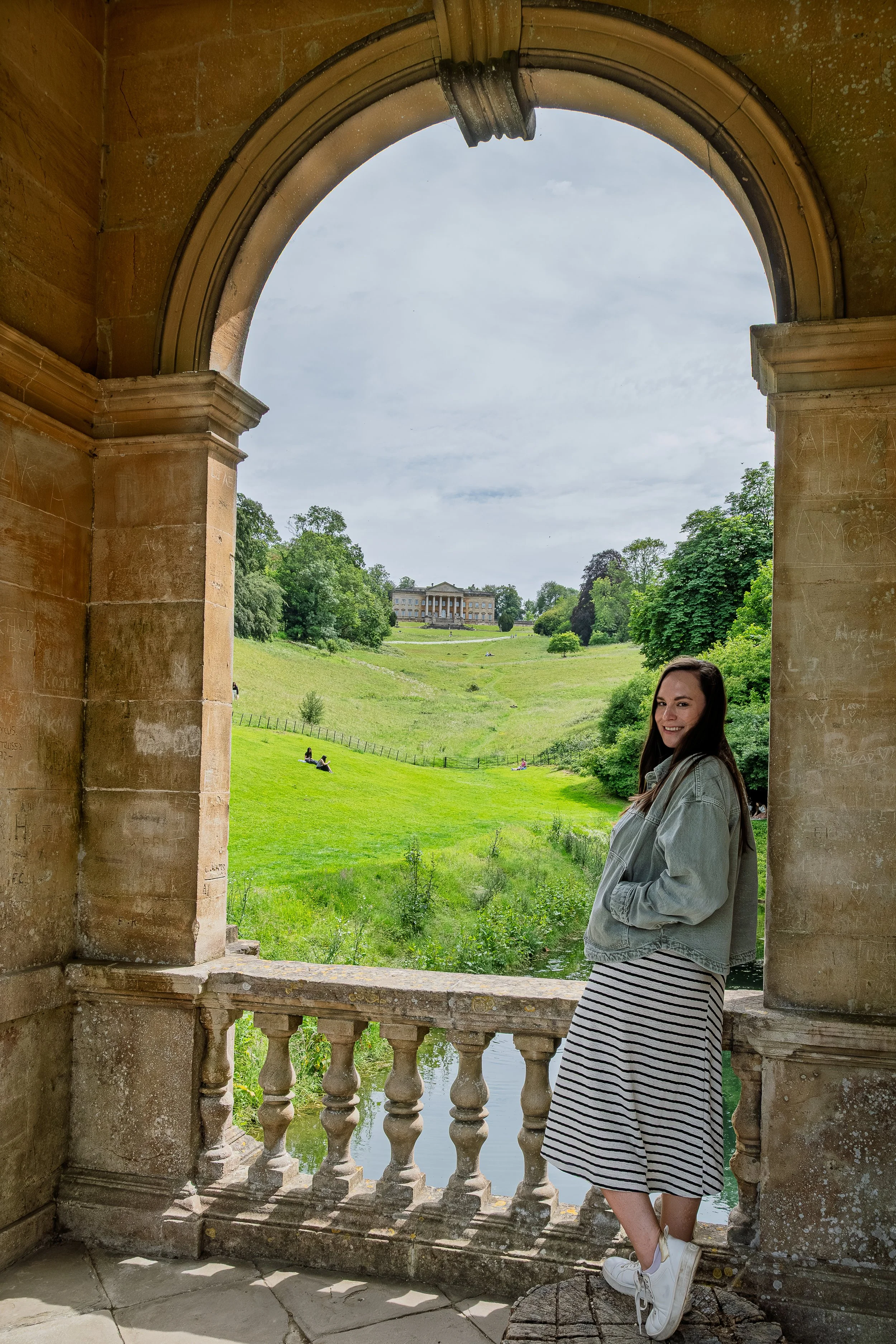 A woman with long brown hair, wearing a gray jacket, a striped skirt, and white sneakers, stands on a small stone ledge under a stone archway, smiling at the camera. Behind her, the scene opens to a lush green landscape with a river, trees, people relaxing on the grass, and a grand building in the distance under a partly cloudy sky.