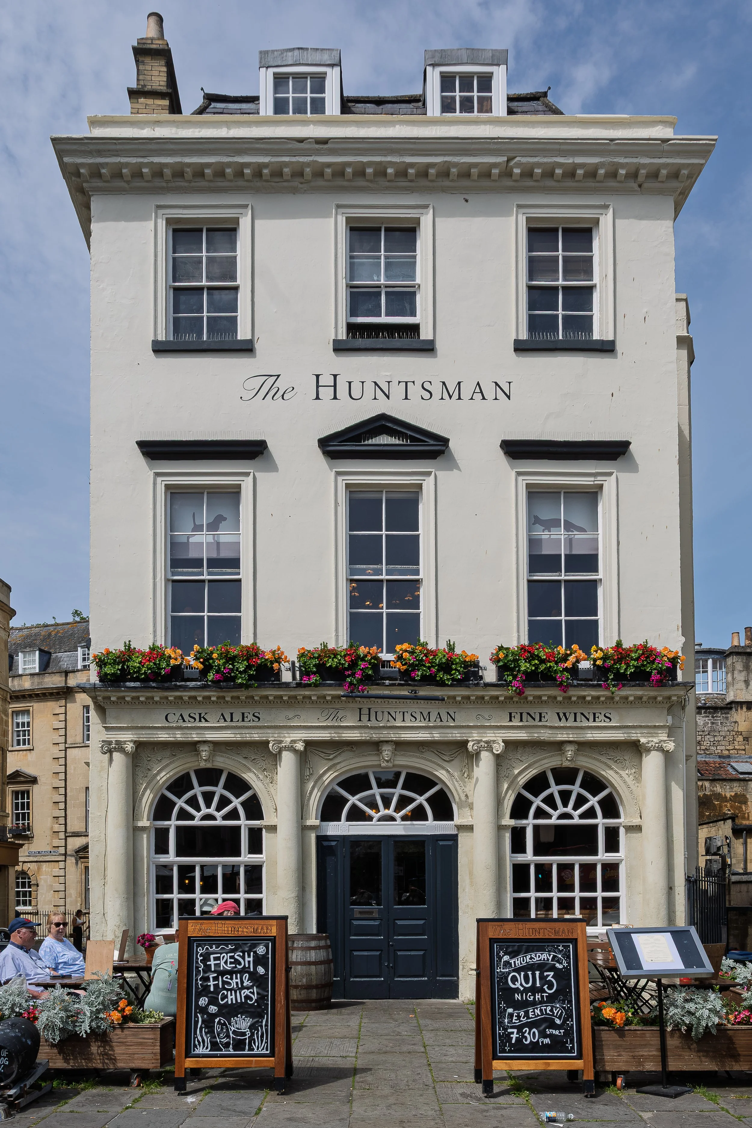 The exterior of a historic pub called The Huntsman, with flowers and outdoor seating. Chalkboards display menus and event information, including fresh fish and chips and a quiz night.