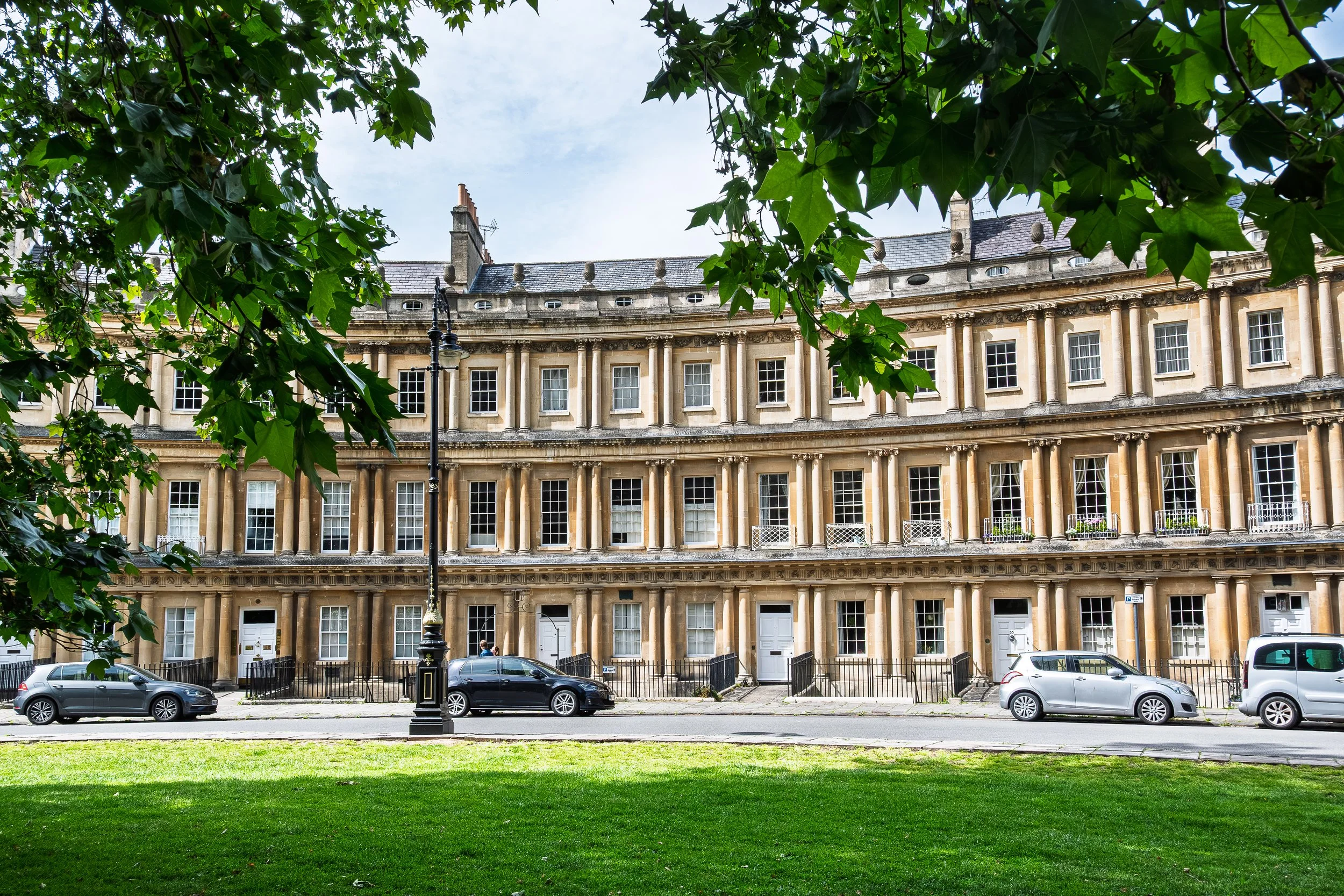 A grand historic building with columns and large windows seen through tree branches with green leaves, parked cars along the street, and a grassy area in the foreground.