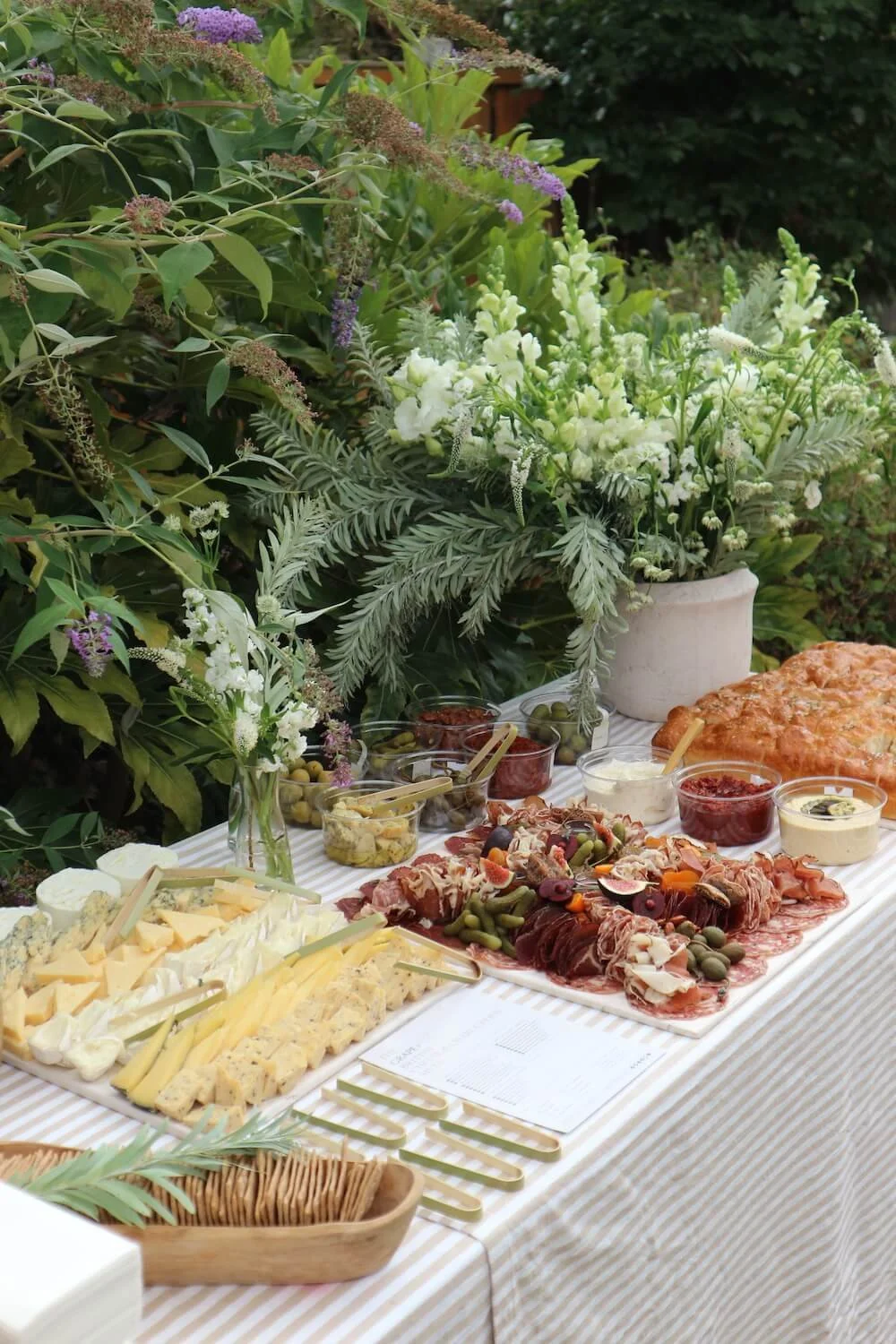 A table set outdoors with an assortment of cheeses, charcuterie, bread, and condiments, decorated with a large arrangement of white and purple flowers and green foliage as part of a garden party or picnic.