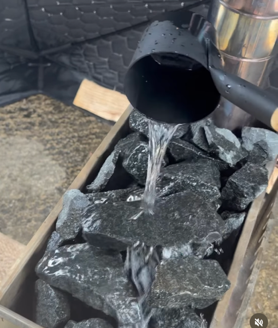 Pouring water into a metal container filled with black rocks or charcoal.