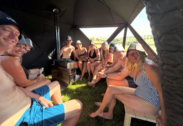 Group of friends sitting inside a tent enjoying a sunny day, some wearing hats and summer clothes, with a small stove in the middle.