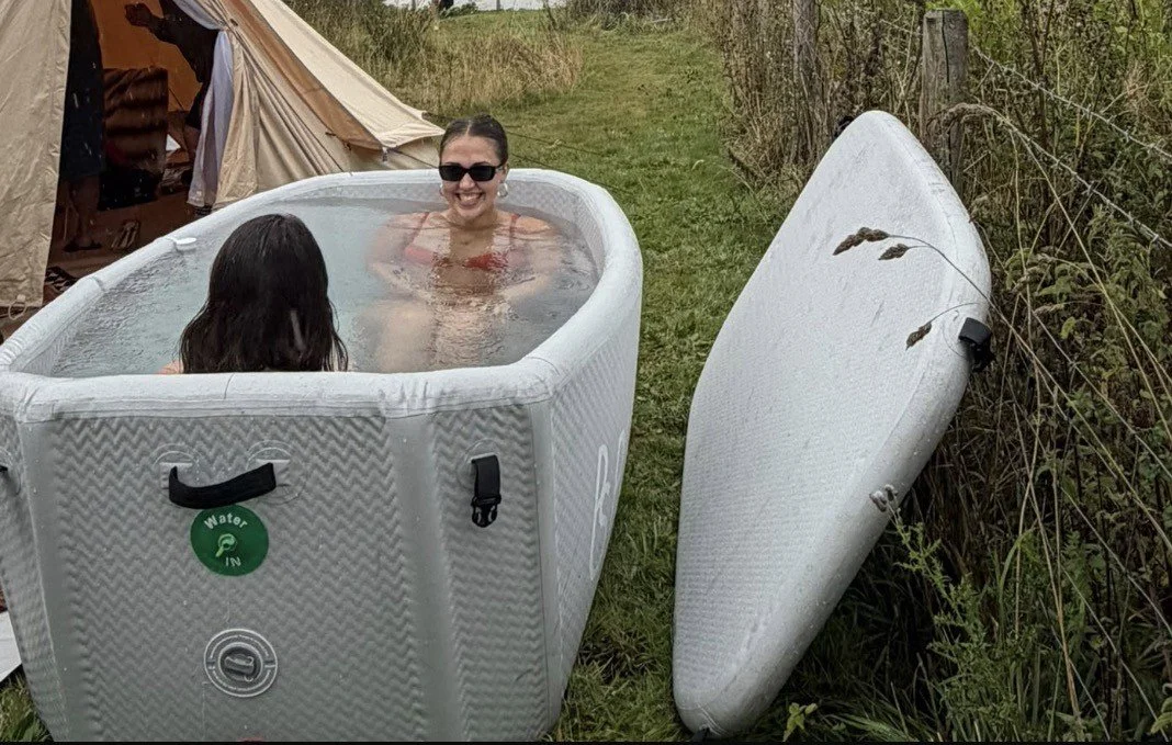 Two women are sitting in an inflatable hot tub outdoors, smiling and wearing sunglasses. One woman has long dark hair, and the other has short hair. A paddleboard rests against tall grass nearby, with a large tent and greenery in the background.