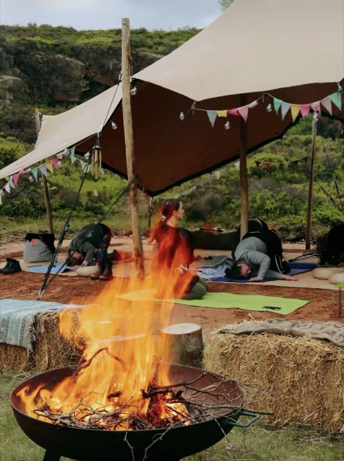 People practicing yoga under a canopy tent outdoors, with a campfire in the foreground, and green hillside in the background.