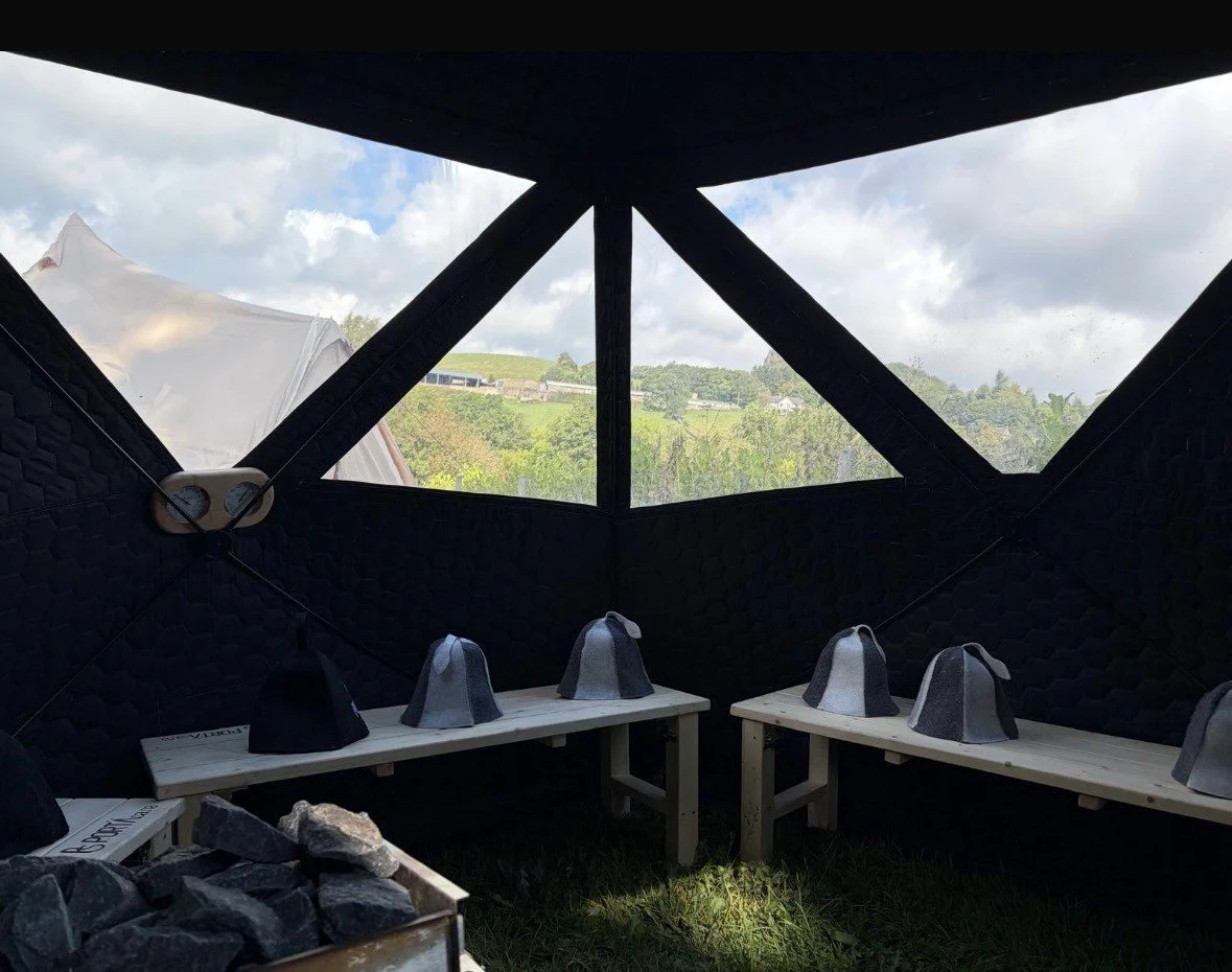 Inside a black yurt with windows showing a green landscape and cloudy sky, two wooden benches with felt hats placed on top of each, and a wood fire pit in the foreground.
