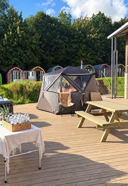 Outdoor scene with a two-tiered tent, a picnic table with bench, and a table with bottled water on a wooden deck, with colorful small cabins and green trees in the background under a partly cloudy sky.