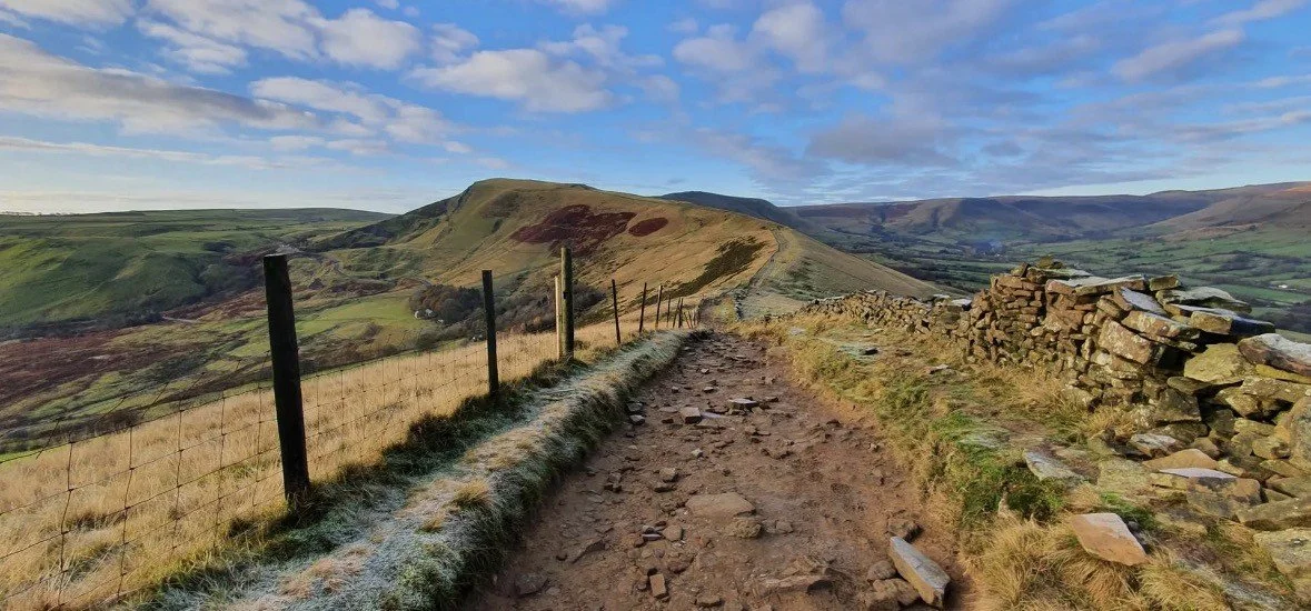 A dirt pathway running along a hillside with a wire fence on the left and a stone wall on the right, leading towards distant rolling hills under a partly cloudy sky.