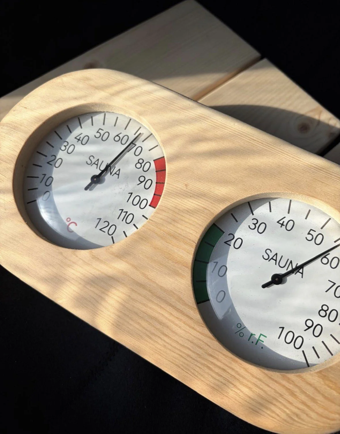 A wooden sauna temperature and humidity monitor with two circular dials. The left dial shows temperature at approximately 60°C, and the right dial shows humidity at around 55%.
