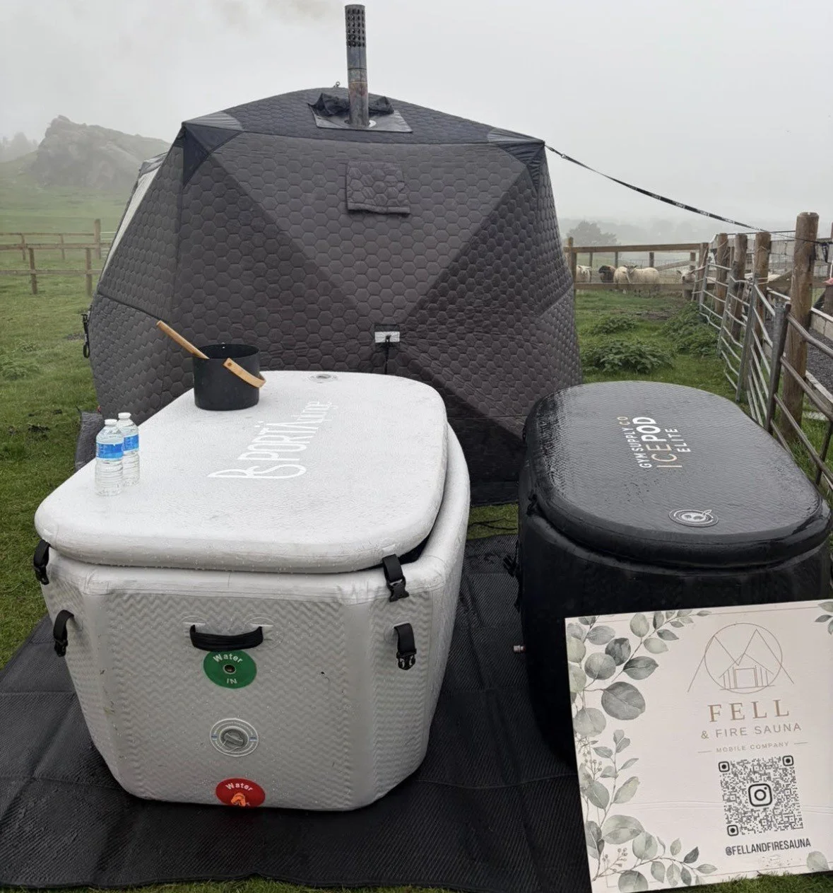 A portable ice bath setup with a white insulated tub, a black insulated tub, and a tent made of black hexagonal insulated panels, set up outdoors in a grassy area with a wooden fence and sheep in the background.