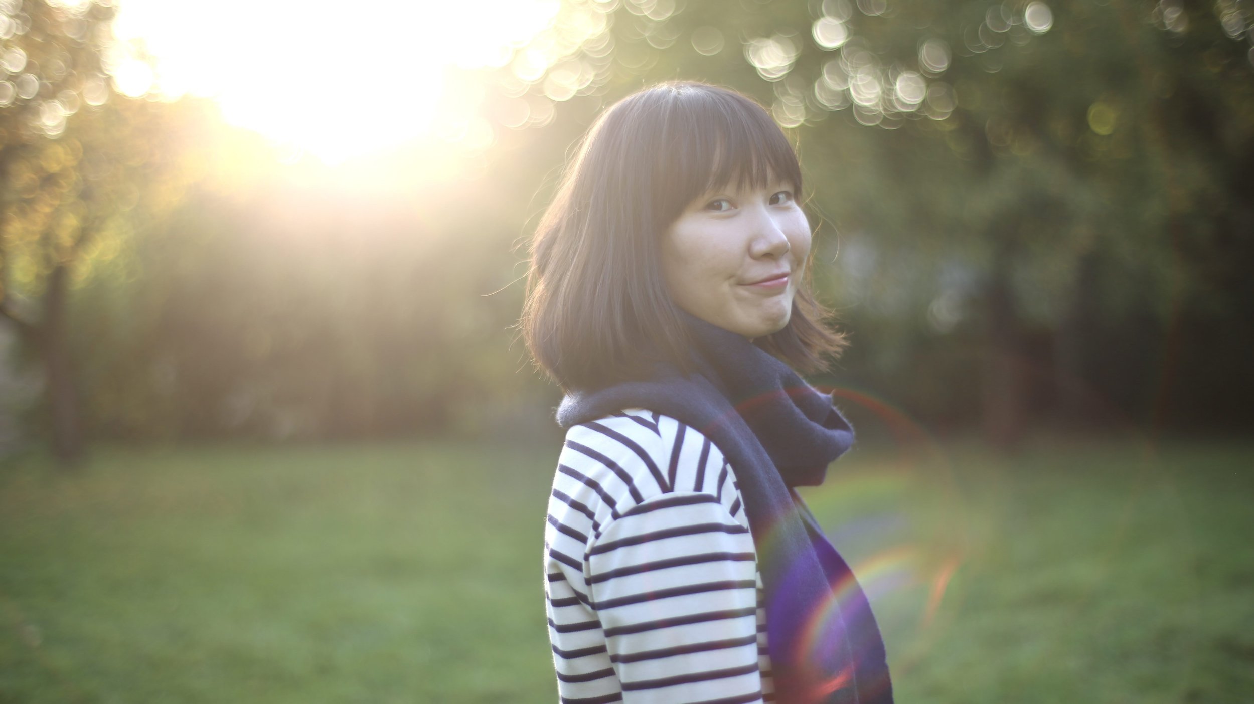 A woman with short brown hair wearing a striped shirt and scarf, standing outdoors in a park during sunset, with trees in the background.
