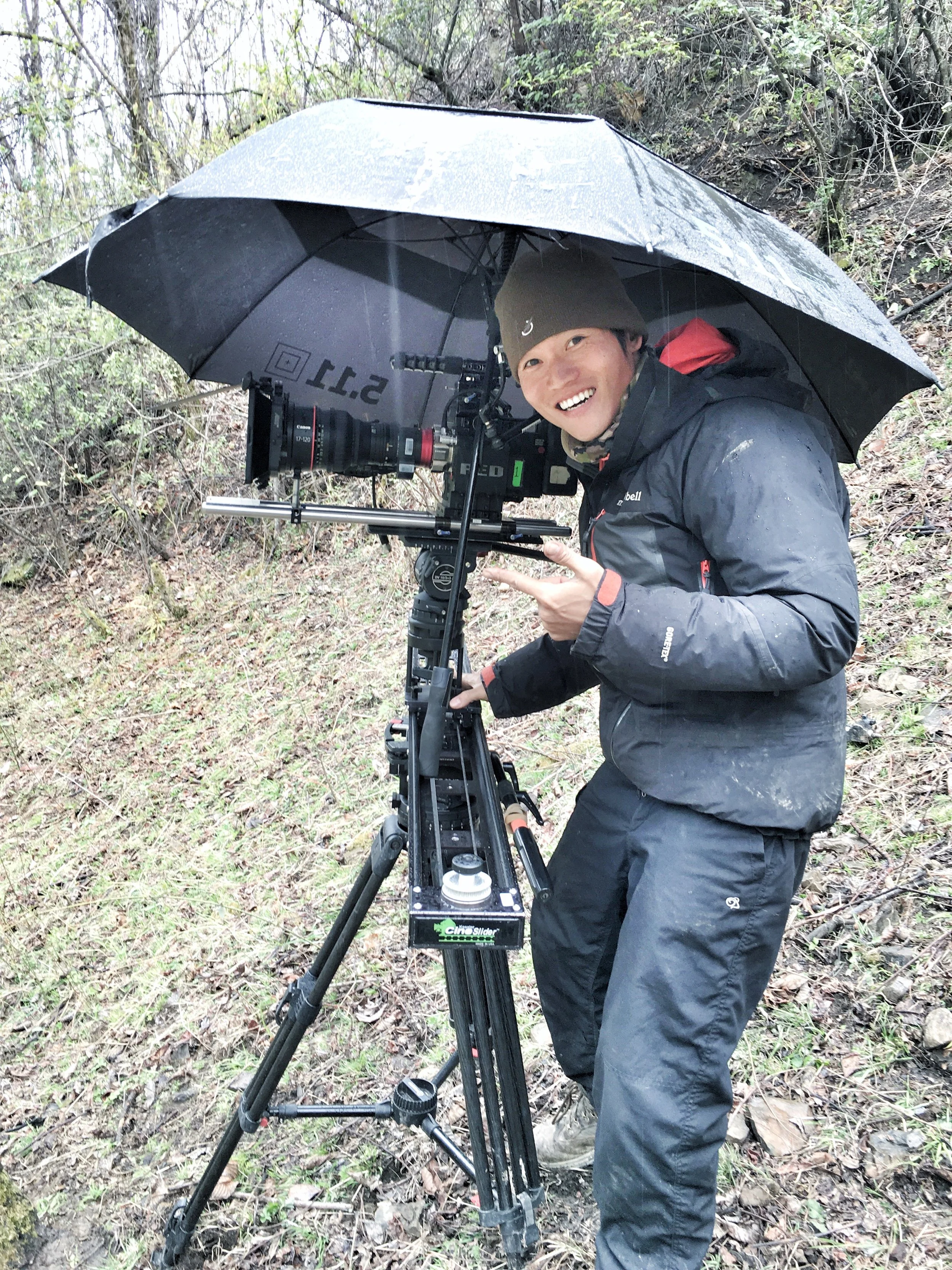 A person smiling and pointing at a camera mounted on a tripod outdoors in a wooded area. The person is wearing a gray beanie, black jacket, and black pants, and is holding an umbrella over the camera.