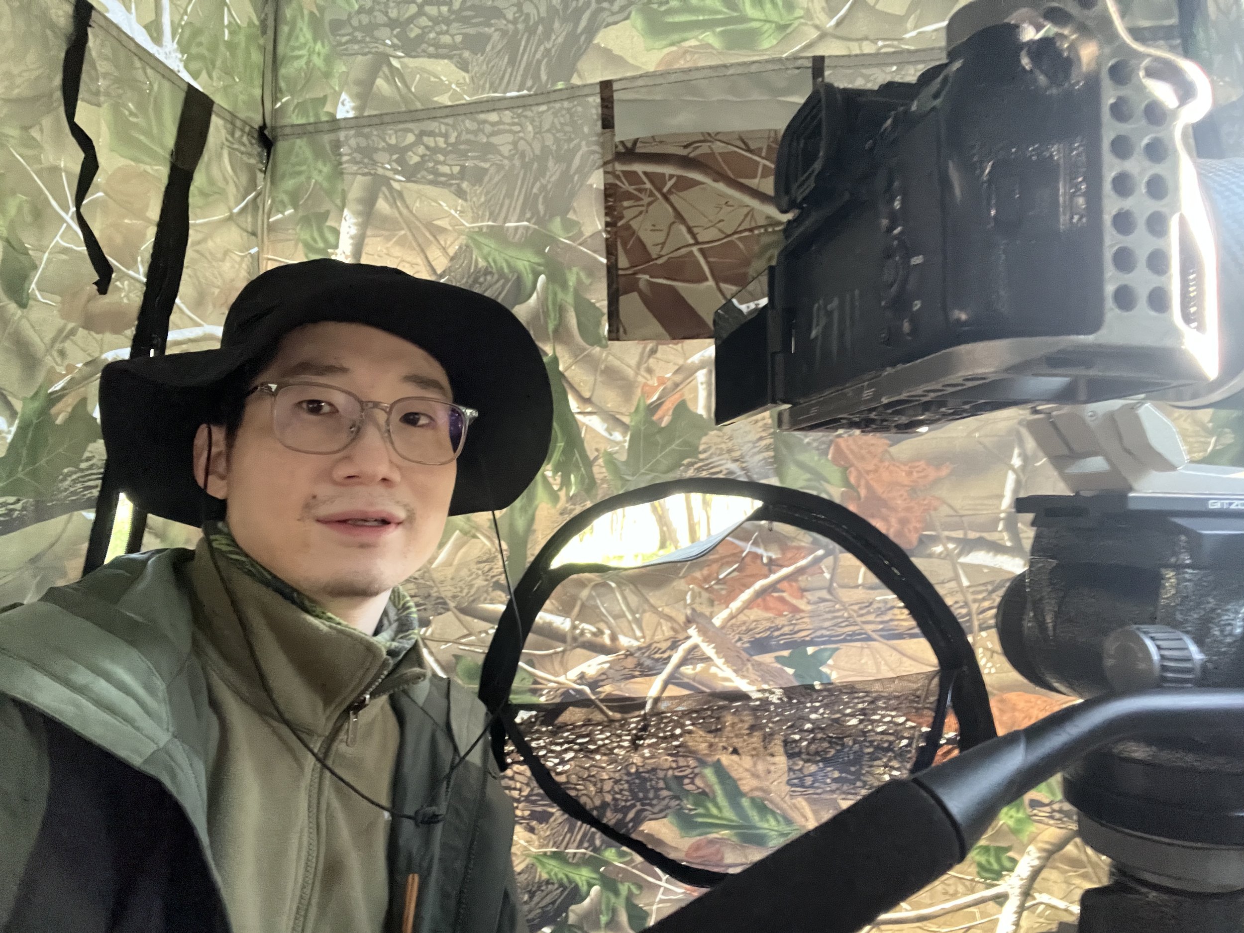 A person with glasses and a wide-brimmed hat posing inside a camouflage hunting tent, with a raccoon trap and wooded environment visible.