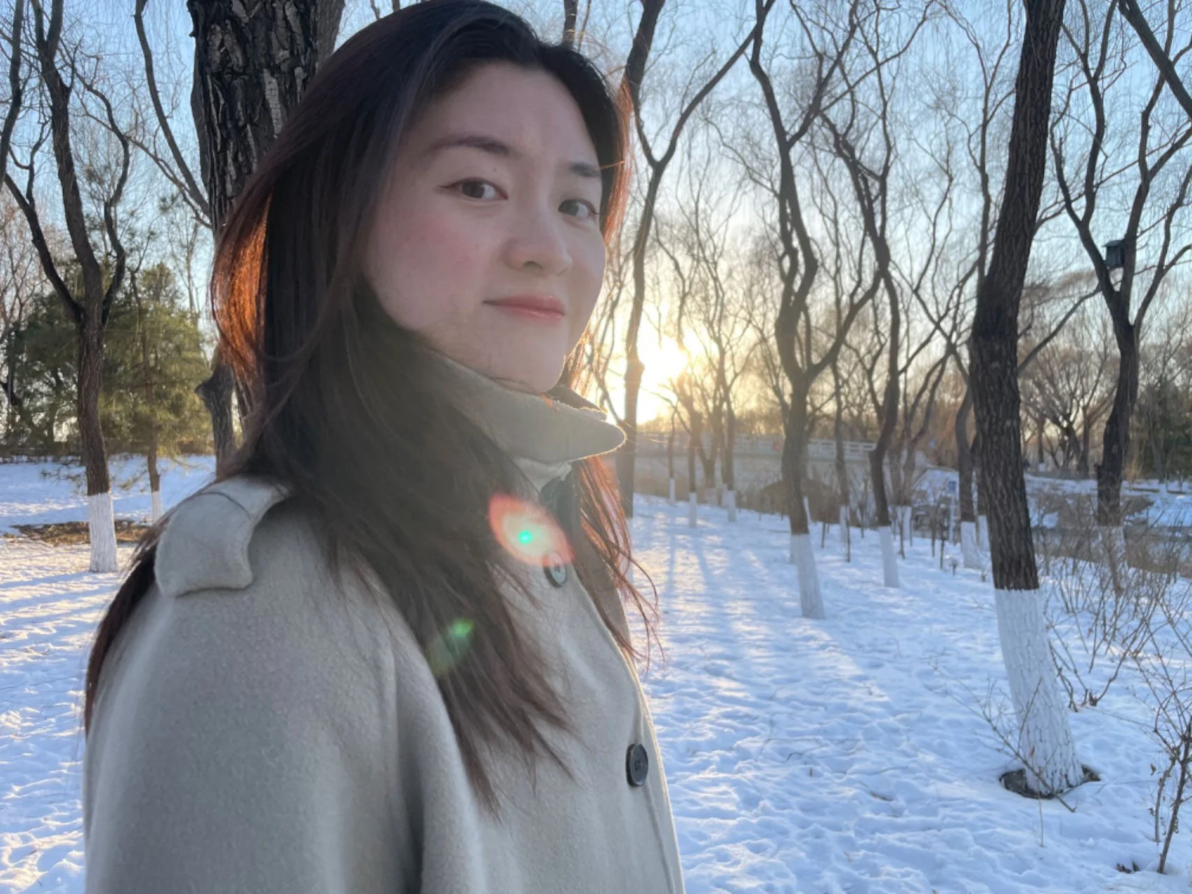 A woman with long dark hair wearing a light-colored coat in a snowy park during sunset.
