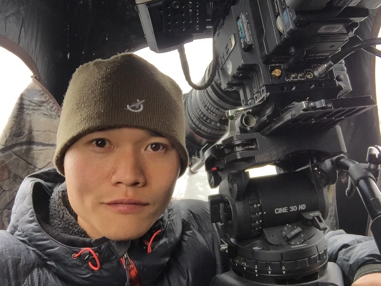 A man wearing a brown beanie and gray jacket taking a selfie inside a camera setup, with professional camera equipment visible.