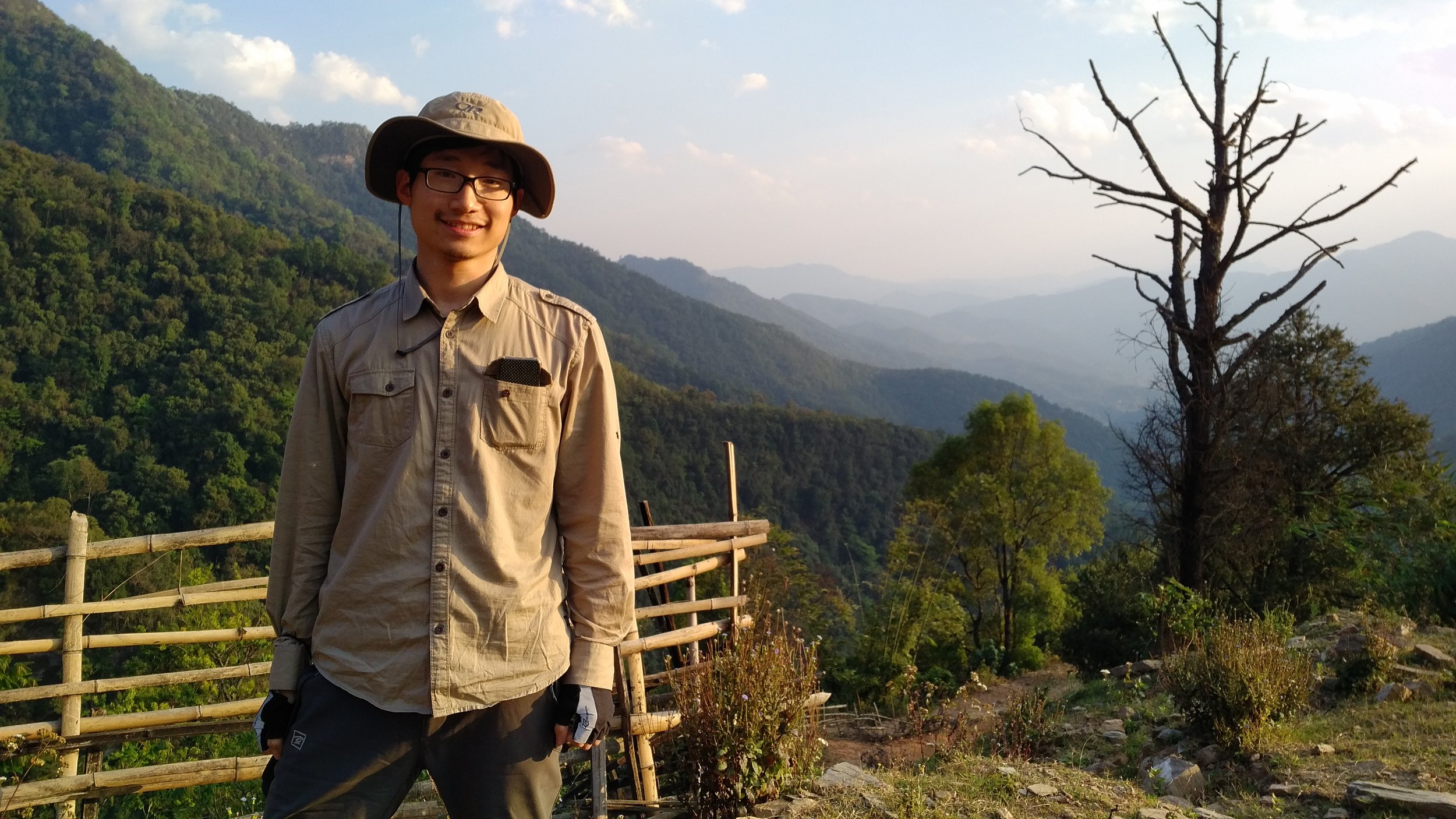 A young man wearing glasses, a wide-brimmed hat, and outdoor clothing standing on a mountain trail with a wooden fence. Behind him are green, forested hills, a dead tree, and a partly cloudy sky.