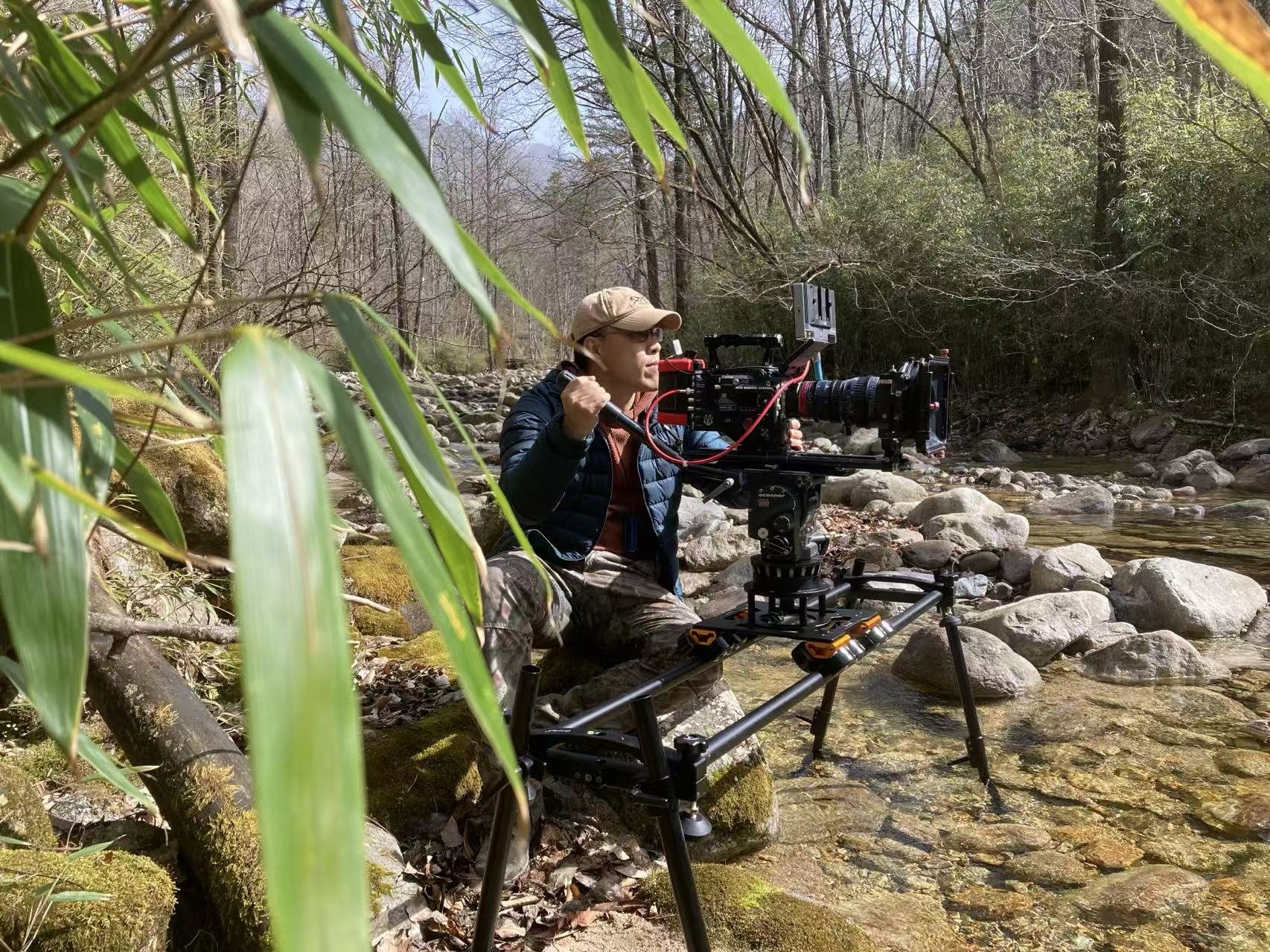 A person operating a professional film camera by a rocky creek in a forest, surrounded by trees and green foliage.