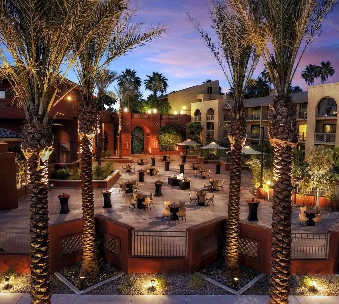 A courtyard with palm trees, outdoor seating, and umbrellas at dusk, surrounded by multi-story buildings with balconies.