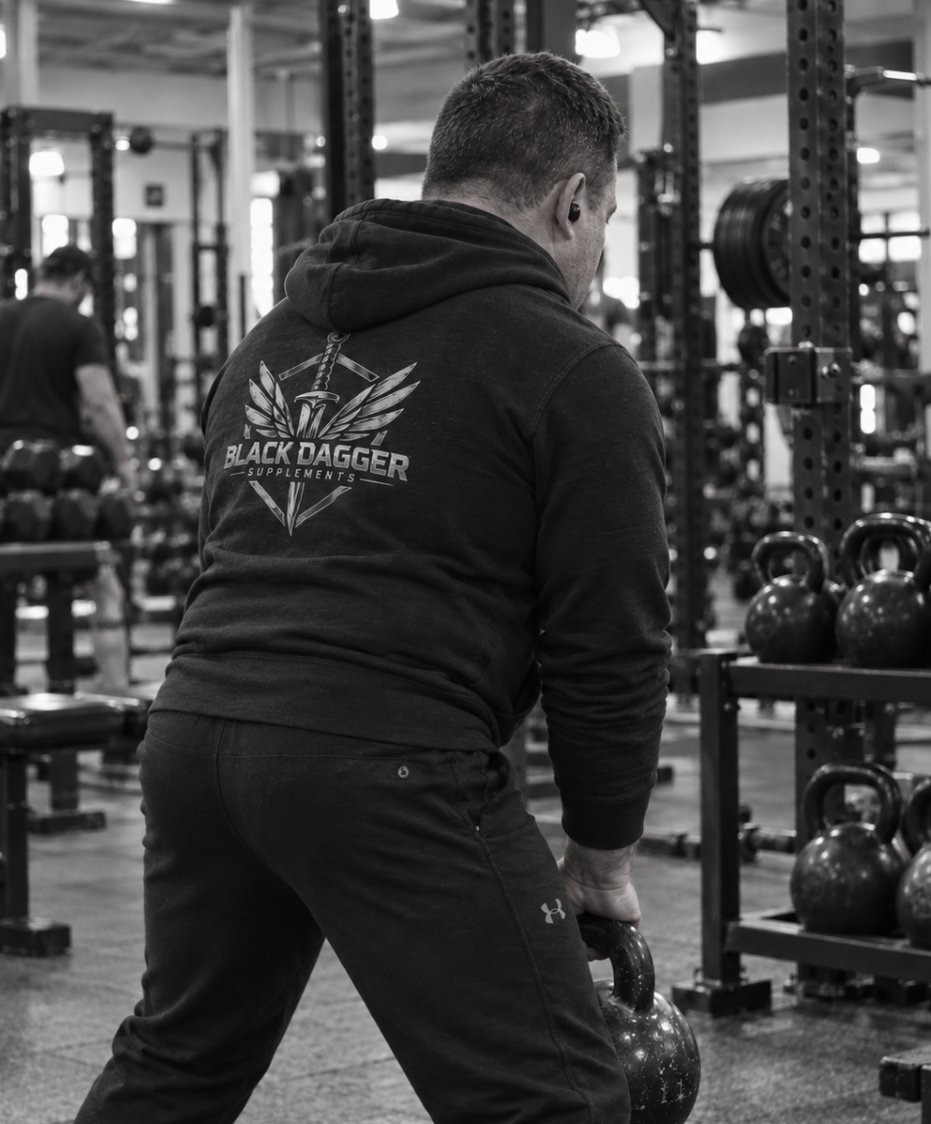 A man performing a kettlebell exercise in a gym, wearing a Black Dagger Supplements hoodie.