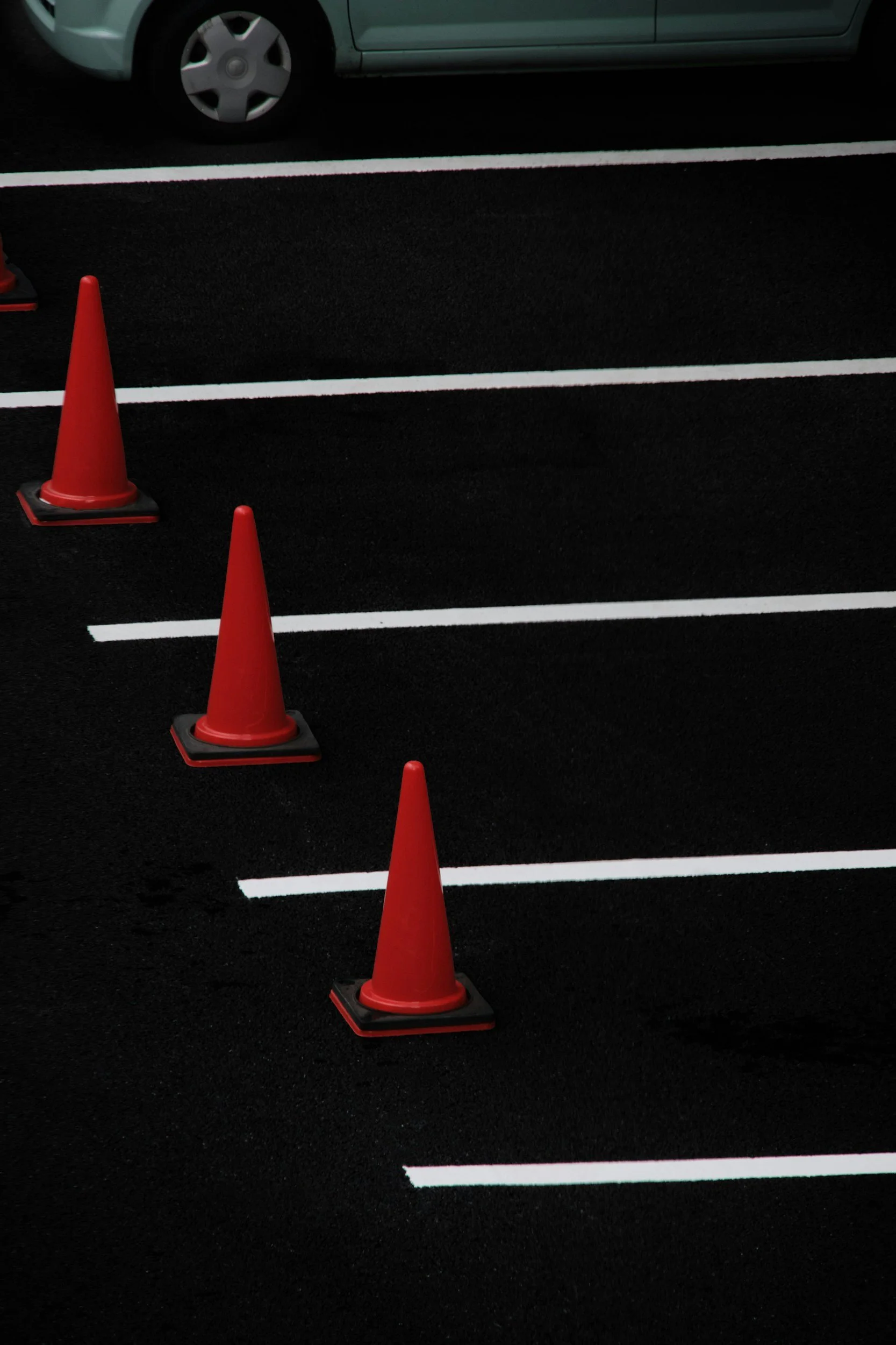 A parking lot with white painted lines and orange traffic cones, with a green car parked in the background.