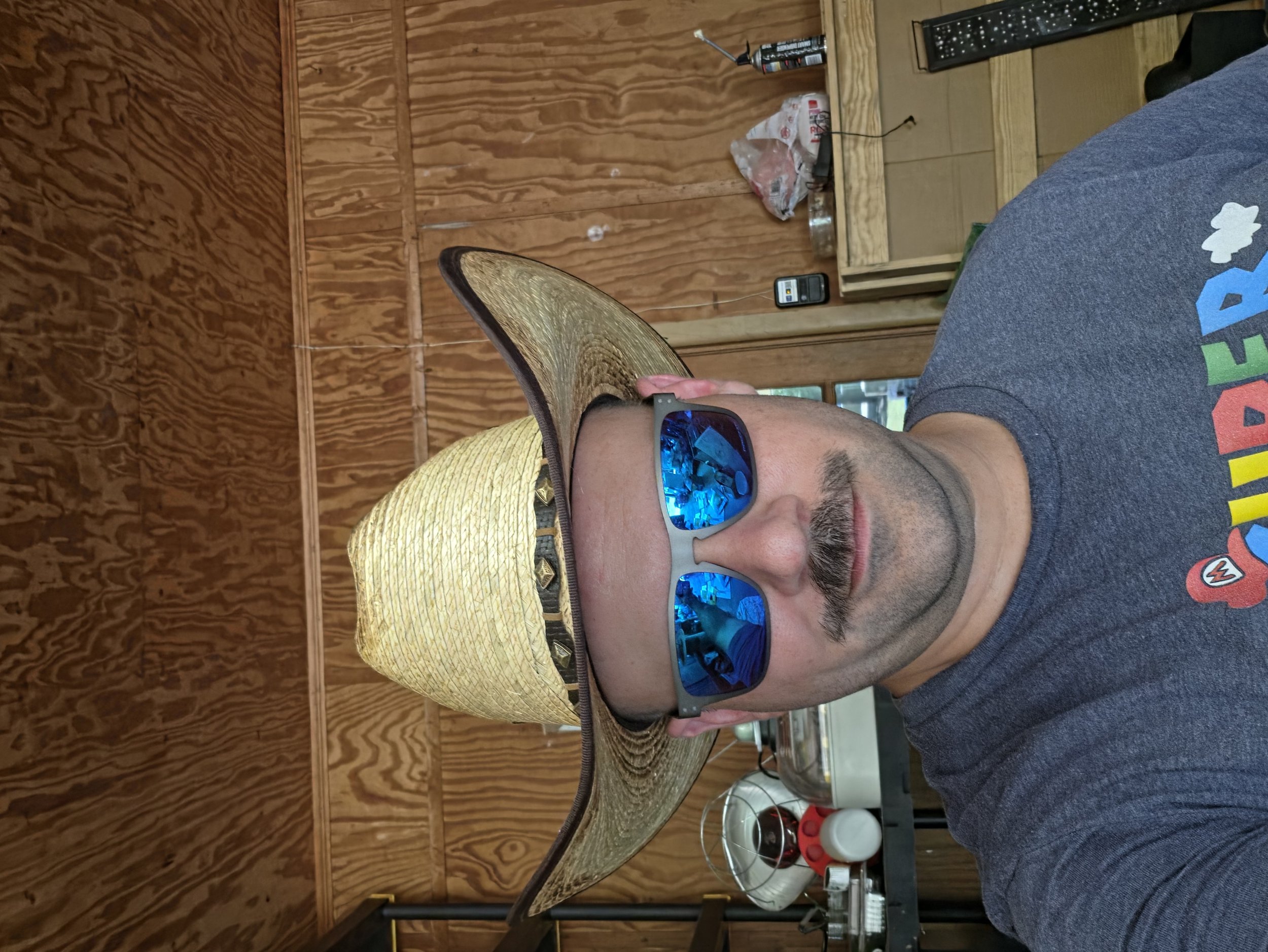 Man wearing sunglasses and a yellow straw hat, partially visible on the right side, in a wooden room with a table and various objects in the background.