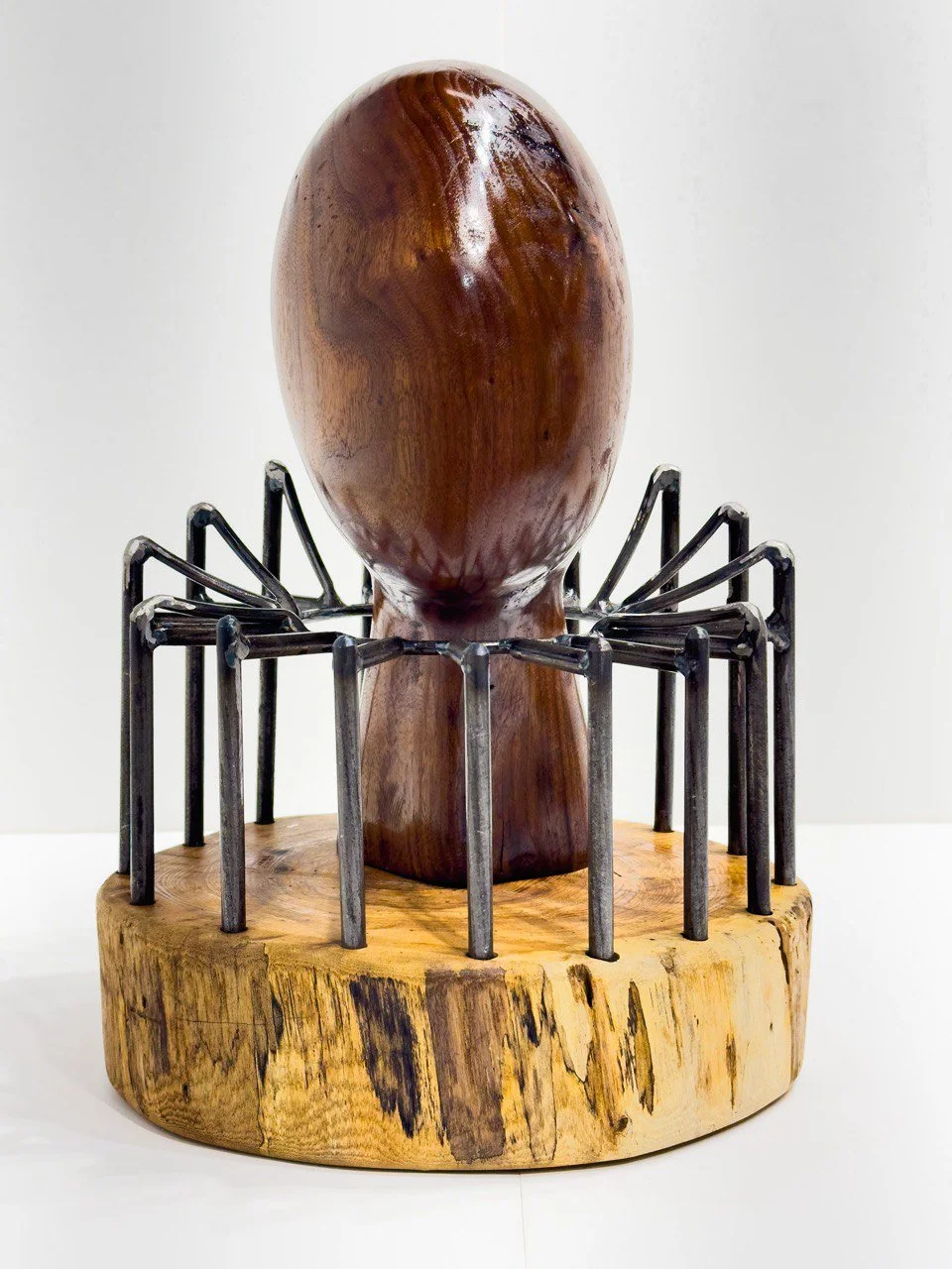 Captive
2023
Wood & metal
12" × 11" × 17.7"

This piece features a wooden head devoid of facial features, mounted on a wooden base and encircled by a series of metal rods that project upward from the base. The absence of facial representation gives t