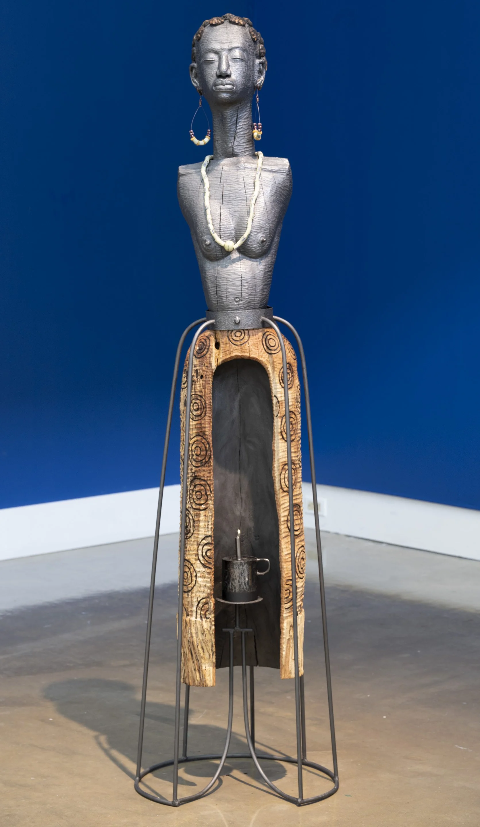 Aku
2024
Wood & metal
18.7" × 17" × 69.5"

This piece depicts a topless female figure intricately carved from wood and adorned with metal earrings, a necklace, and a skirt that is carefully hollowed out from the front. Blending sculptural craftsmansh