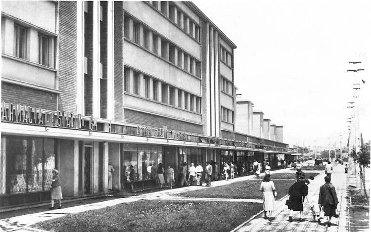 Black and white photo of a busy city street with pedestrians walking along the sidewalk in front of a multi-story building with storefronts.