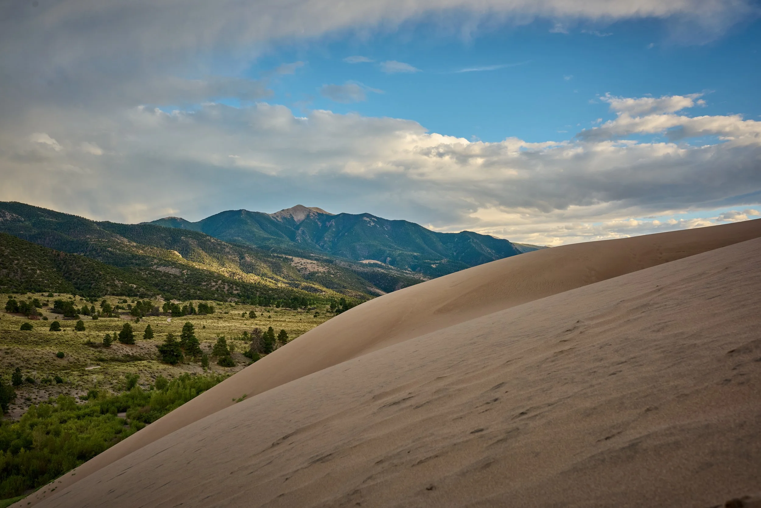 Sand dunes in the foreground with green mountains and trees in the background under a partly cloudy sky.