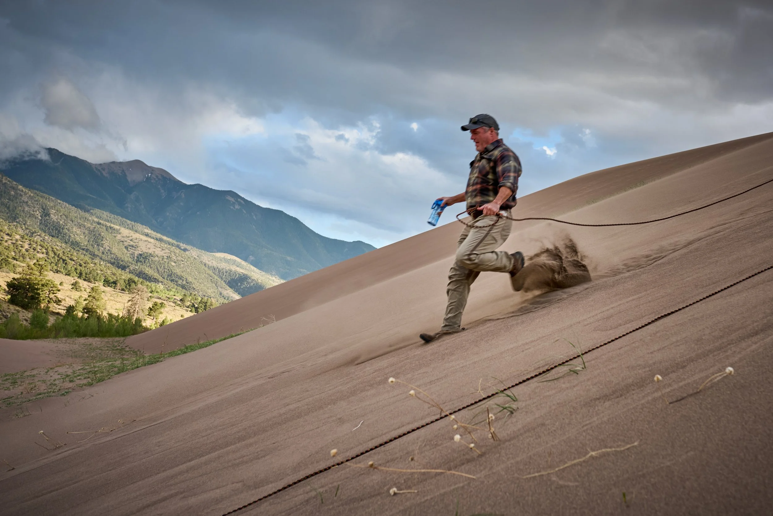 A man in outdoor clothing holding a water bottle and moving quickly down a sandy slope with a mountain landscape in the background.