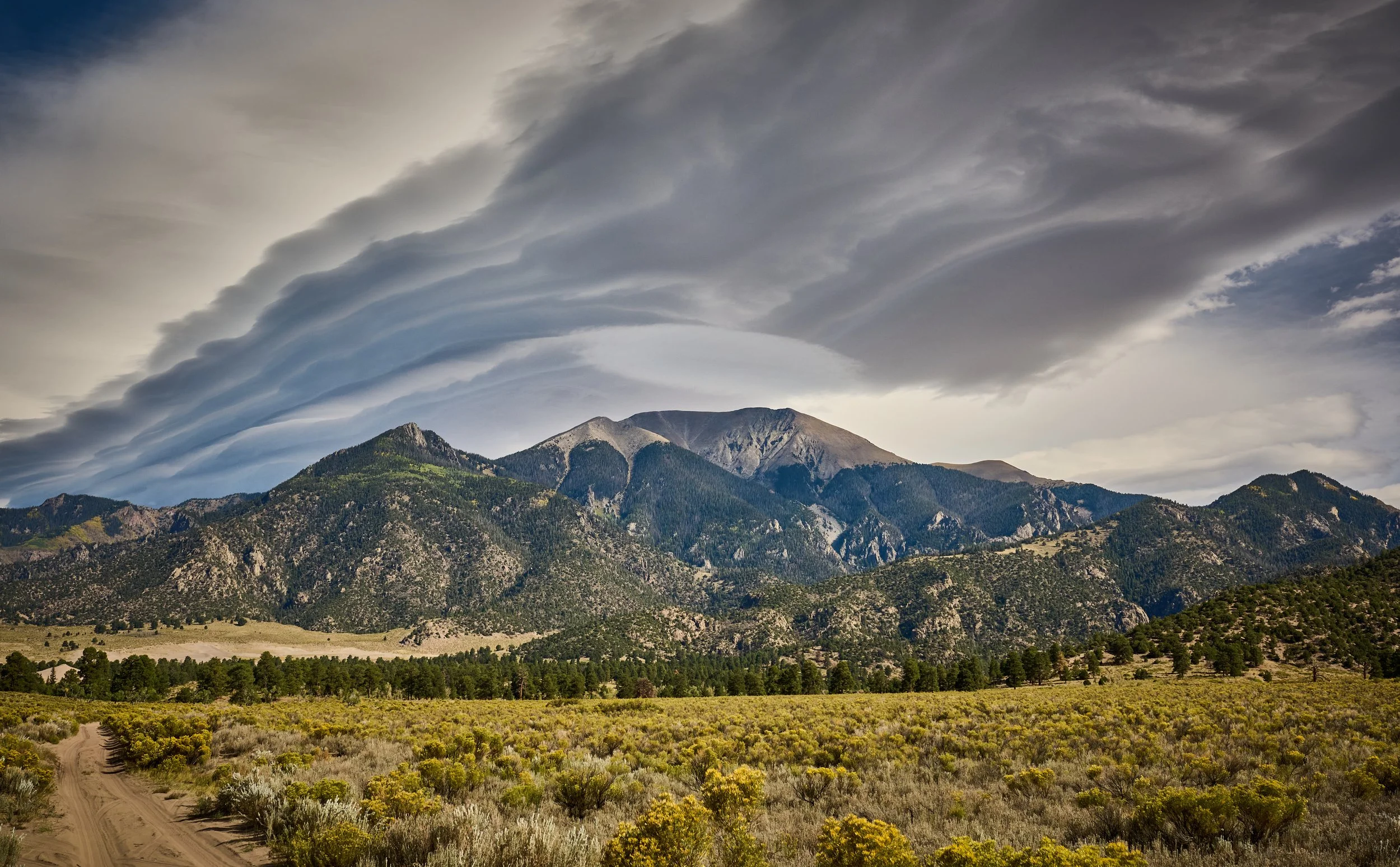 A mountain landscape with clouds overhead, a dirt trail, and green and yellow shrubs in the foreground.