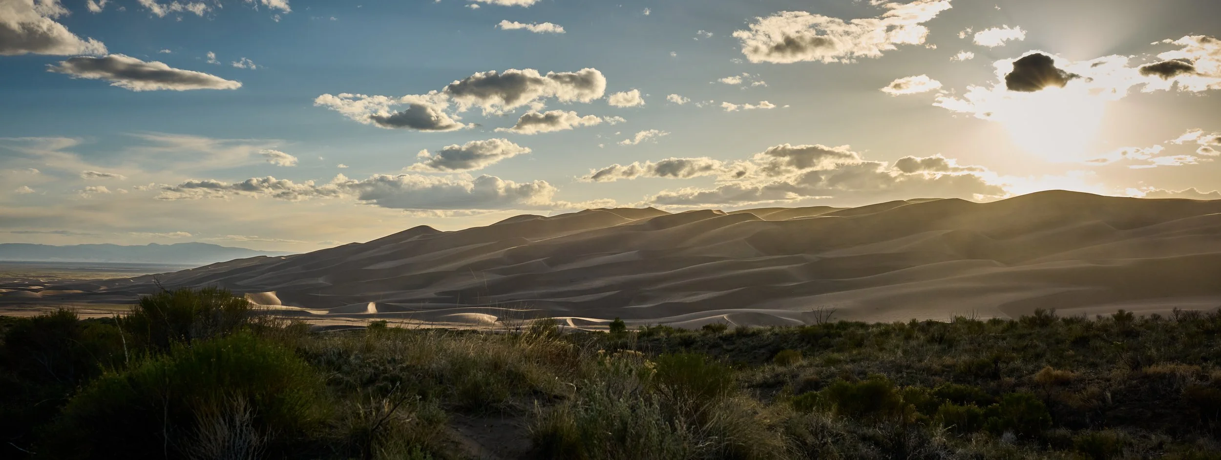 Sunset over sand dunes with scattered clouds and desert vegetation in foreground.