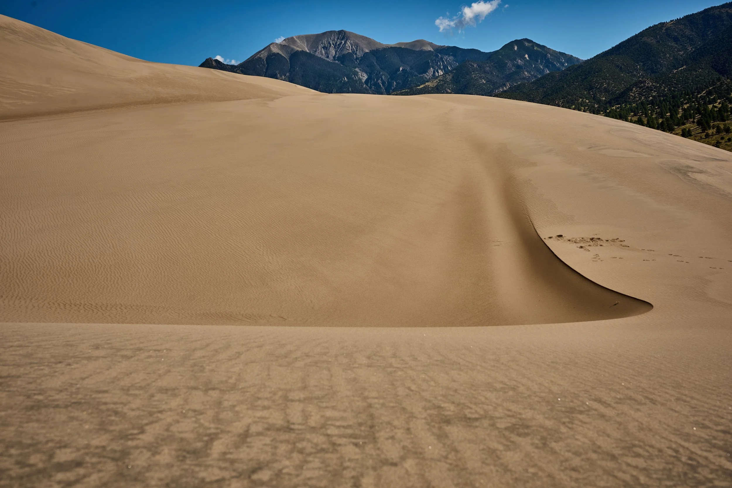 Close-up view of a sand dune with ripples, with a mountain range and blue sky in the background.