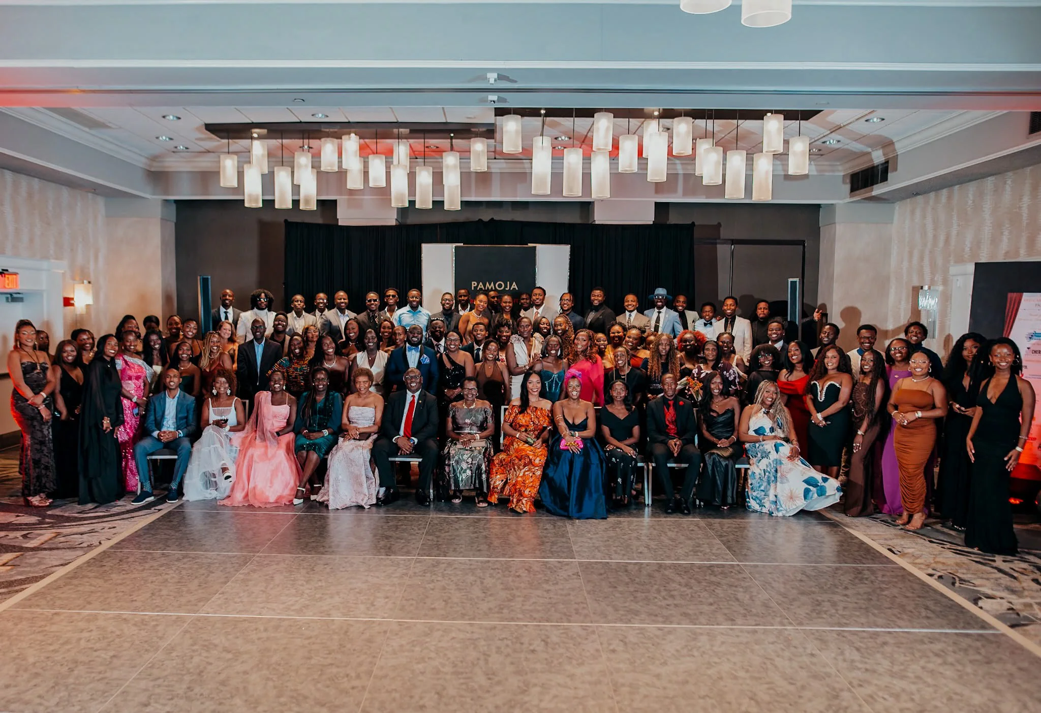 Large group of men and women in formal attire posing for a photo in a conference hall. Some are standing, while others are seated in front. The background features a dark curtain with a sign that says 'PAMOJA' and modern overhead lighting.