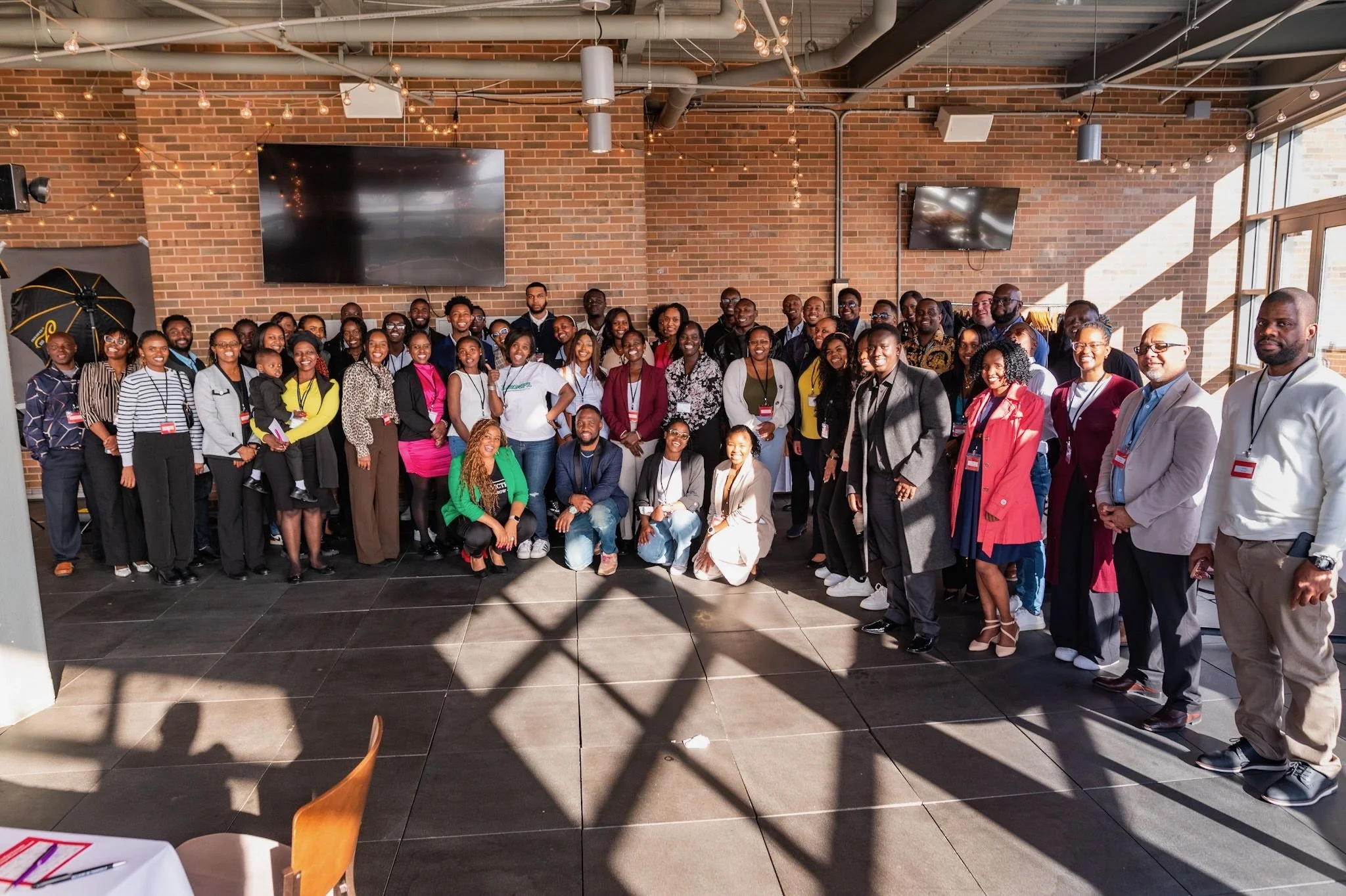 A large group of diverse people posing together indoors with brick walls and large windows, some wearing conference badges.