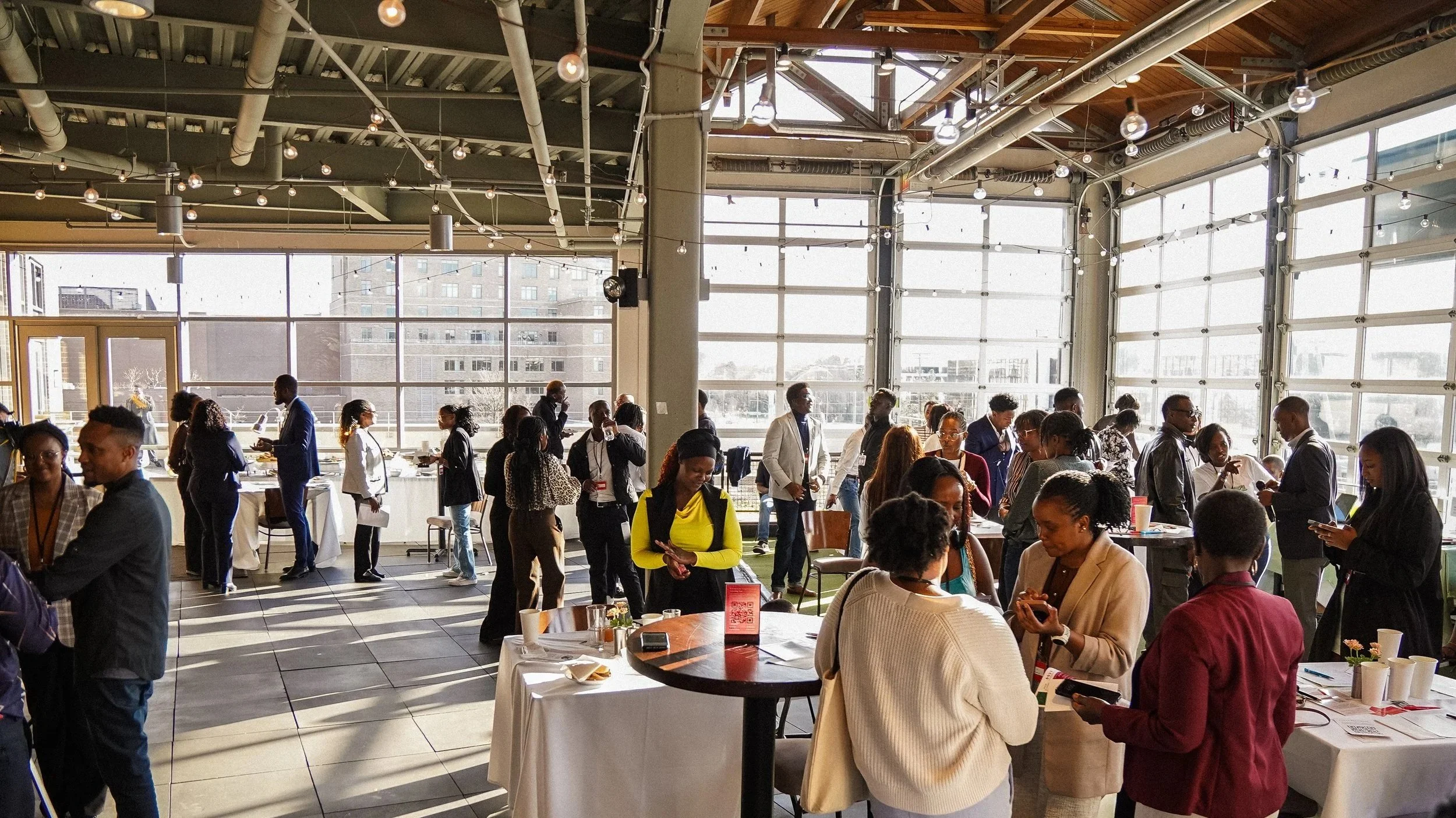 People gathered at a social or professional event in a spacious room with large windows and modern industrial decor, including string lights and exposed pipes.