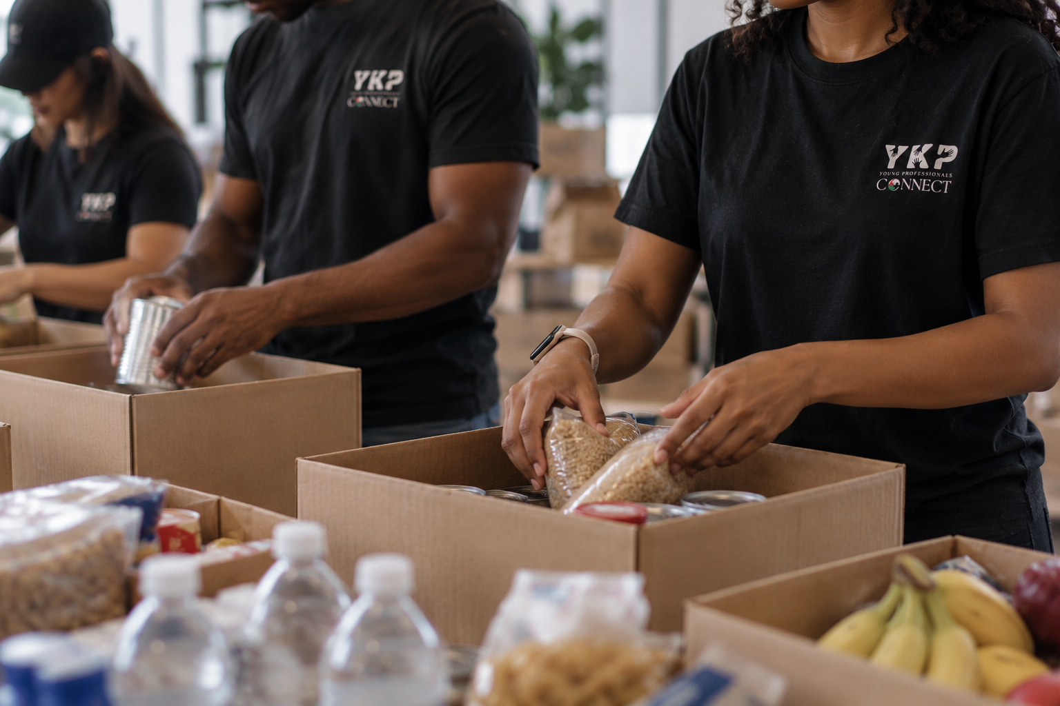 People packing food items into boxes, with some canned goods, bananas, and bottled water visible.