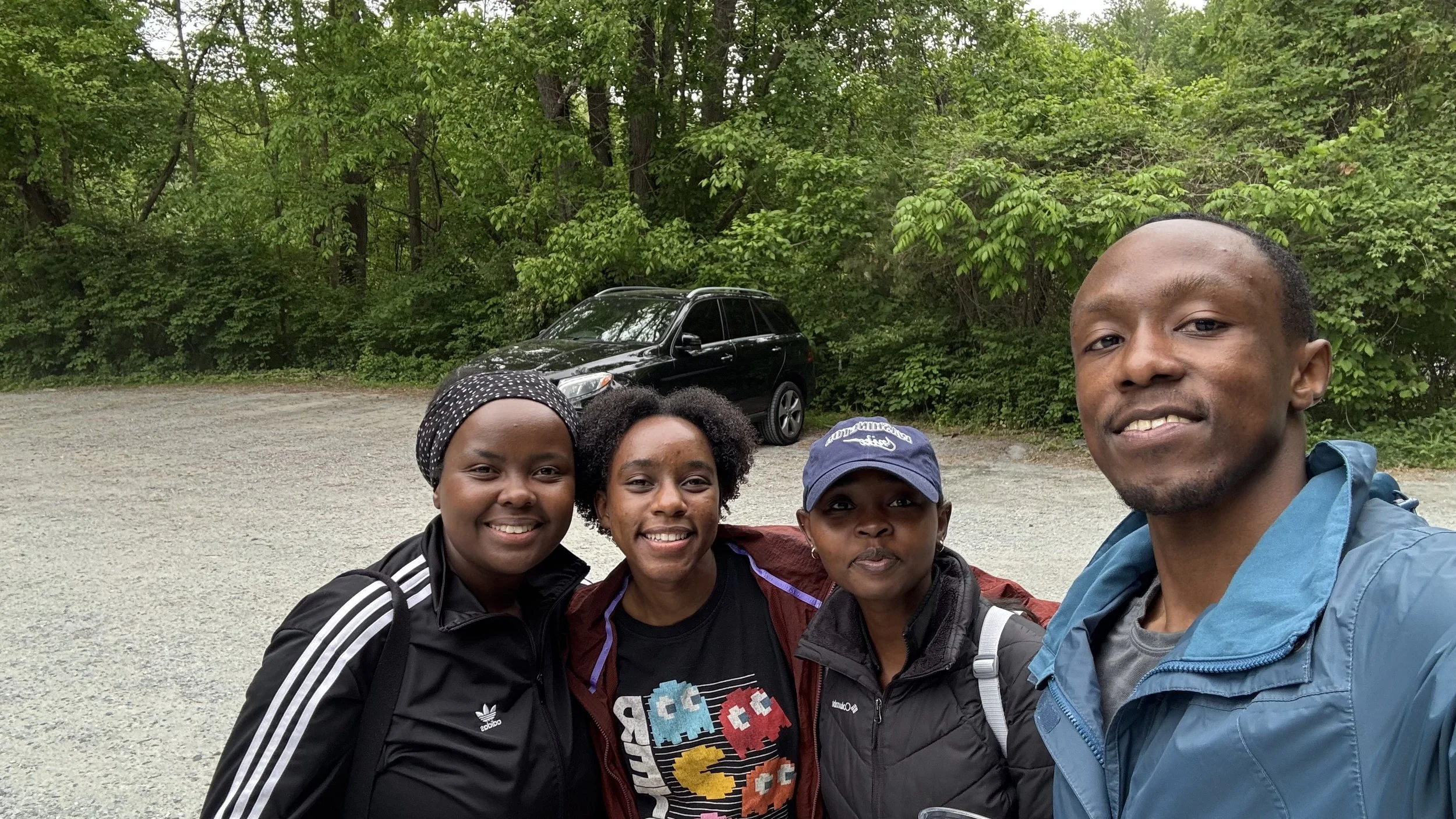 Four smiling people taking a selfie outdoors near a black car and green trees.