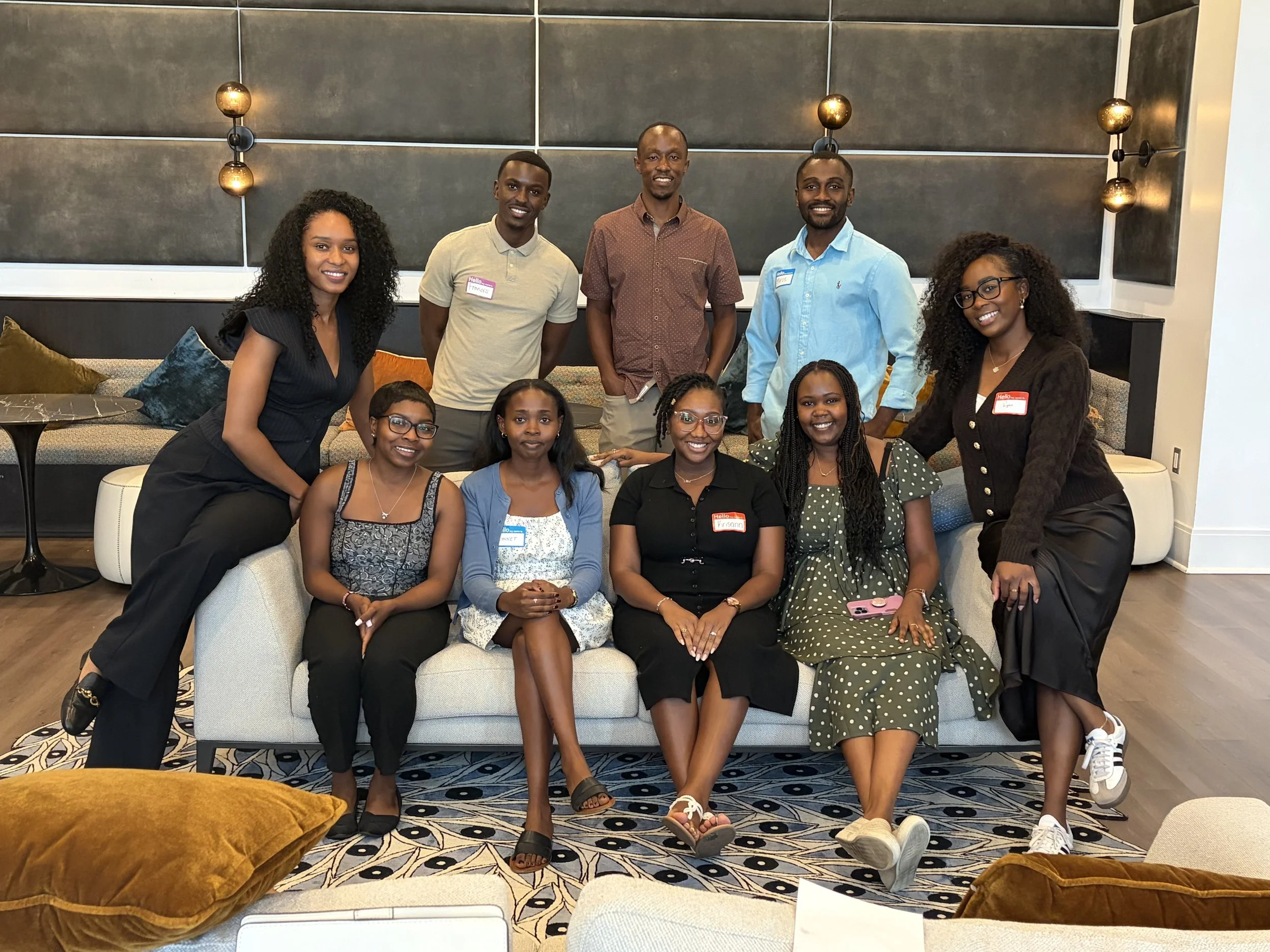 A group of nine diverse people, five women and four men, gathered together in a modern indoor setting. Some are seated on a couch or chairs, while others are standing behind them, smiling at the camera. The background features a wall with modern light fixtures and decorative pillows on the seating area.