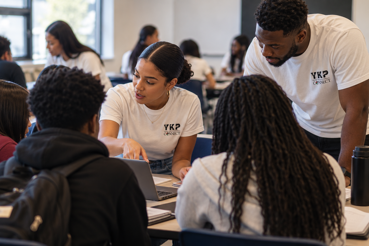 Young adults working together at a table in a classroom or workshop setting, discussing and using a laptop.