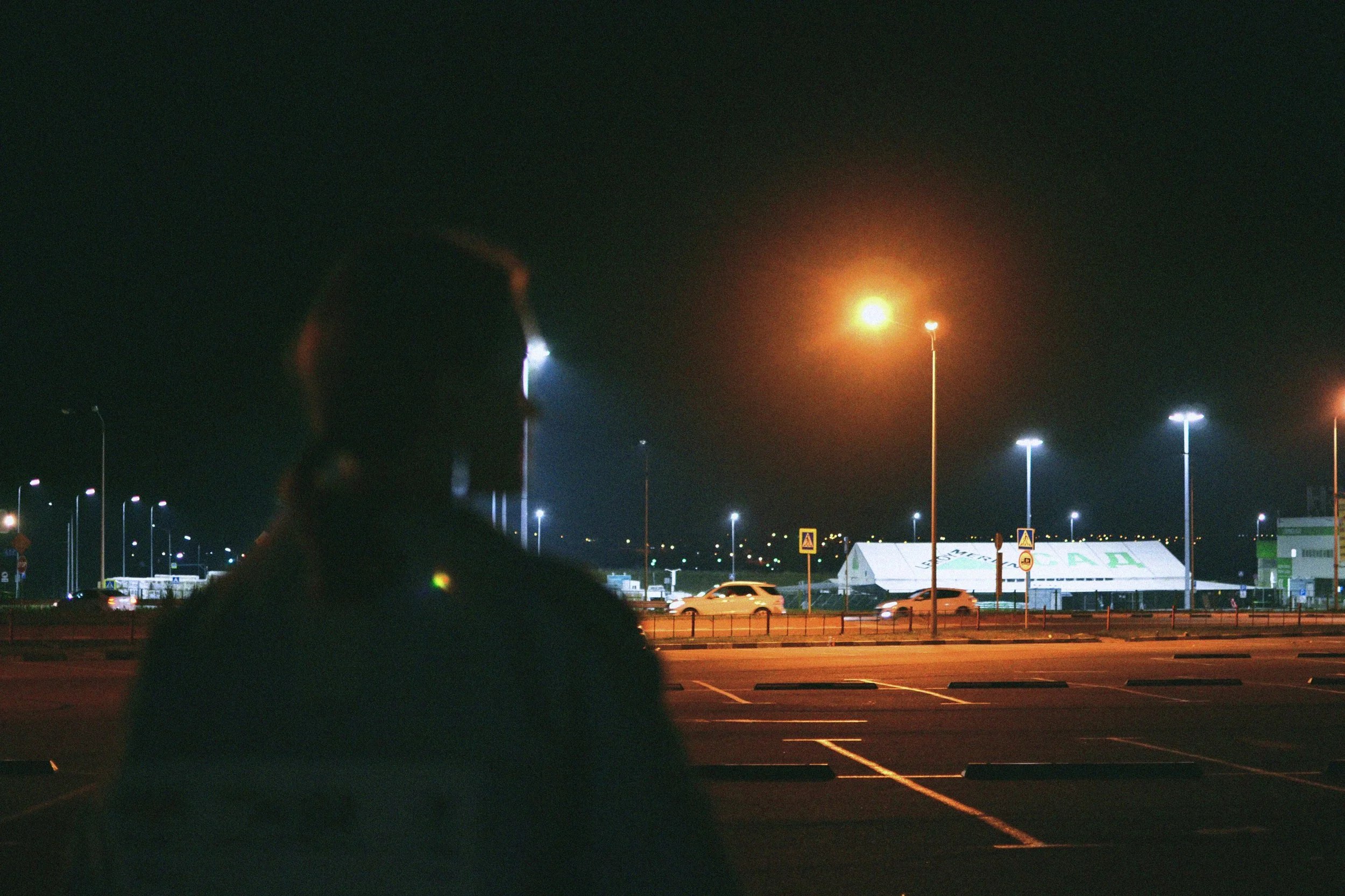 Nighttime scene showing a parking lot with a silhouette of a person's head and shoulders in the foreground. In the background, tall streetlights illuminate the area, with other cars and a large white tent or structure visible.