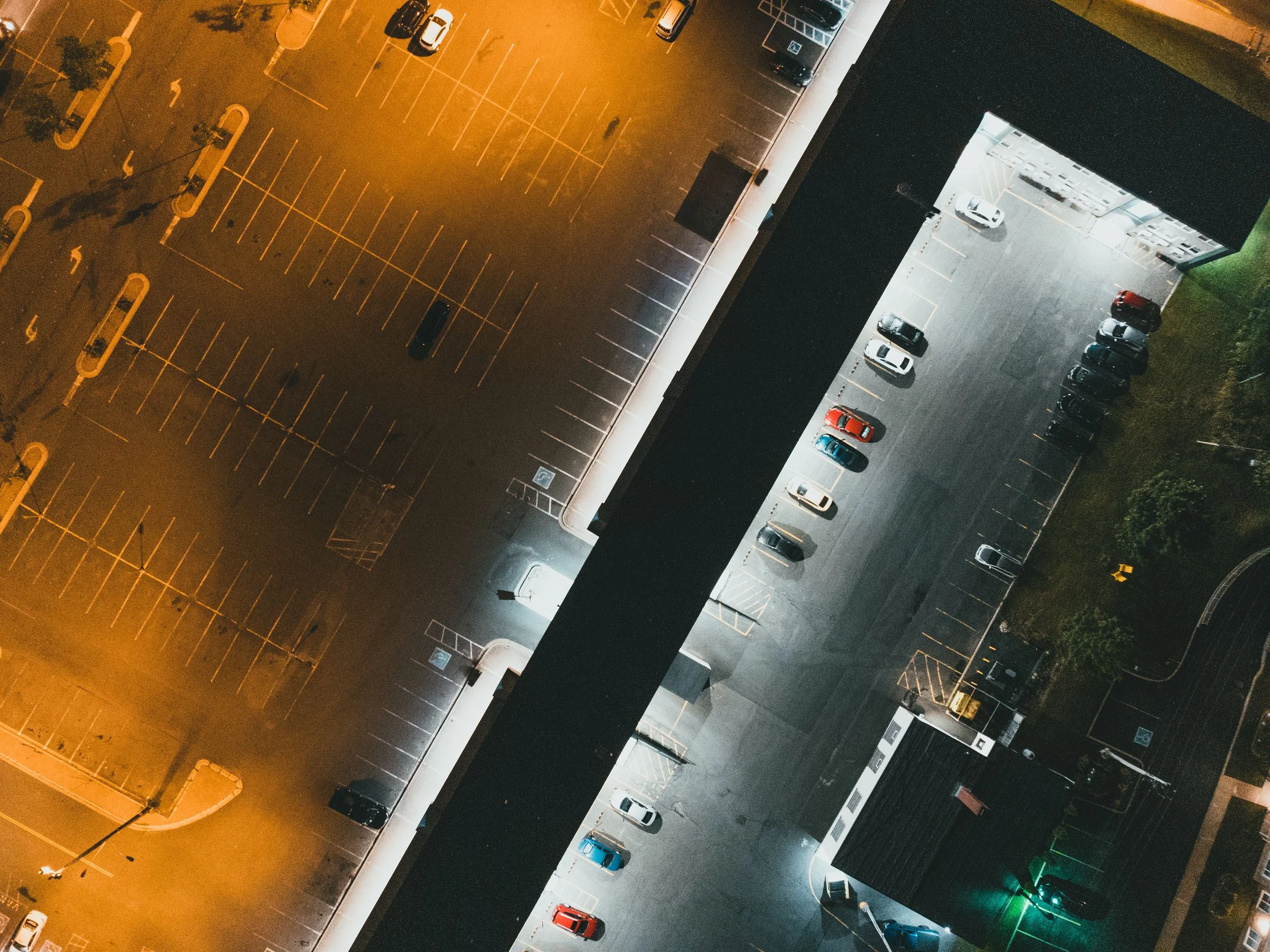 Bird's eye view of an empty parking lot at night with scattered cars and illuminated from streetlights and nearby building lights.