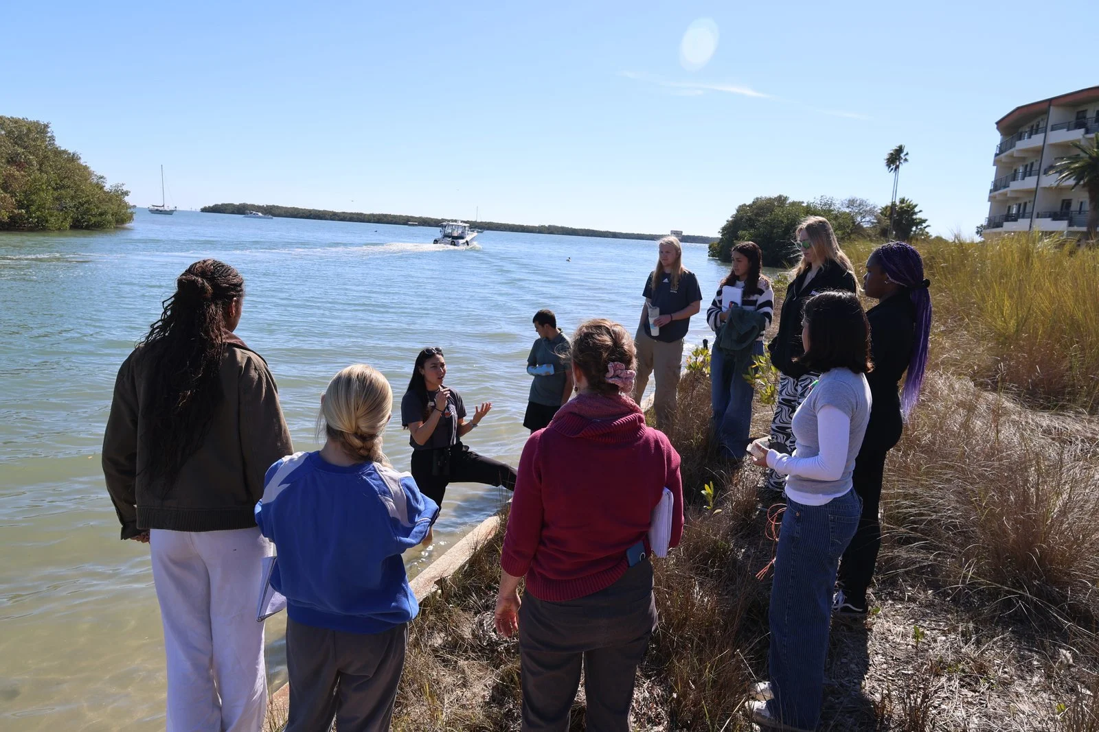 A group of people standing on a beach near the water, listening to a woman who is talking and gesturing with her hand. Several boats are visible on the water, and the sky is clear with few clouds.