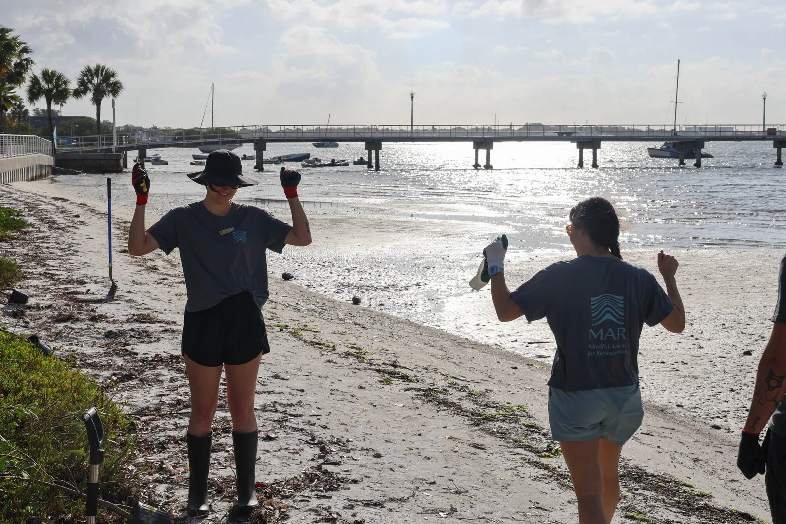 Two women celebrating on a beach near a pier, holding tools, with boats in the water and palm trees in the background.