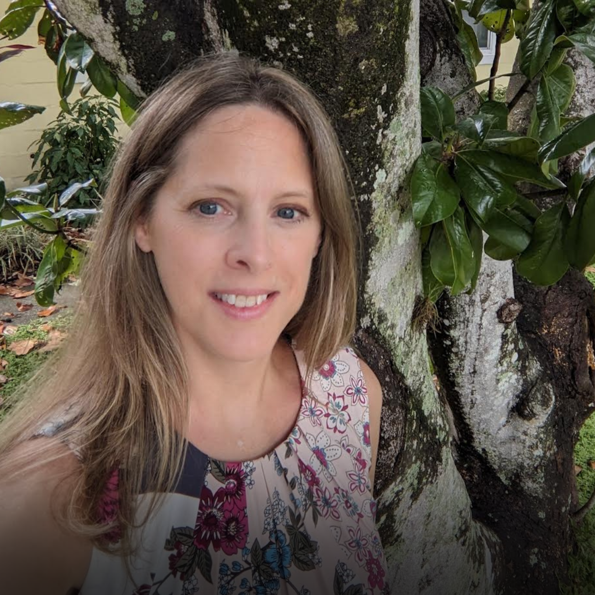 A woman with long light brown hair and blue eyes taking a selfie outdoors next to a tree with dark green leaves.