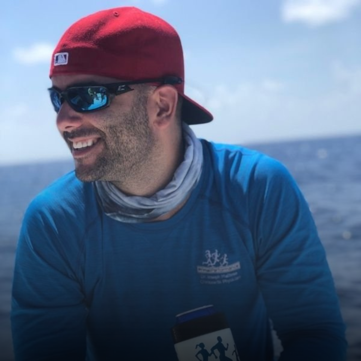 Man wearing a red backward cap, sunglasses, and a blue long sleeve shirt, smiling while sitting outdoors near the water.